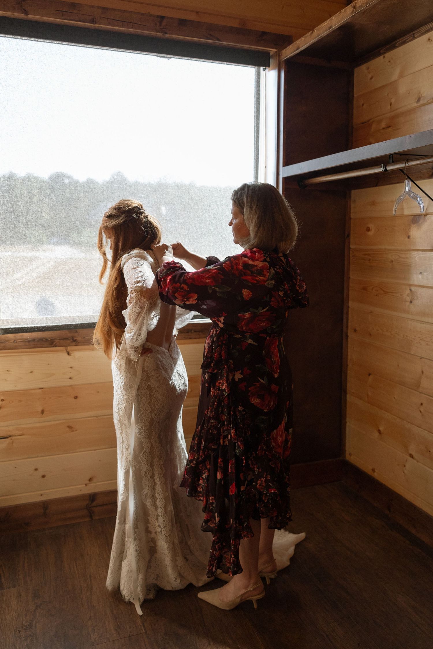 A wedding dress being adjusted in a wooden-paneled dressing room with natural window light.