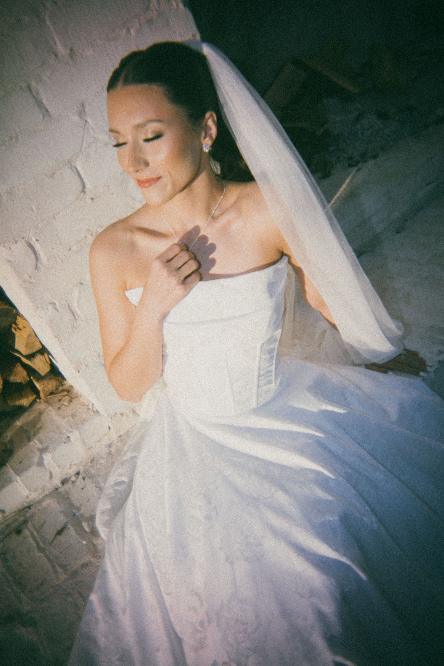 A bride in a white strapless wedding gown and veil stands against a textured wall in vintage-style photography.