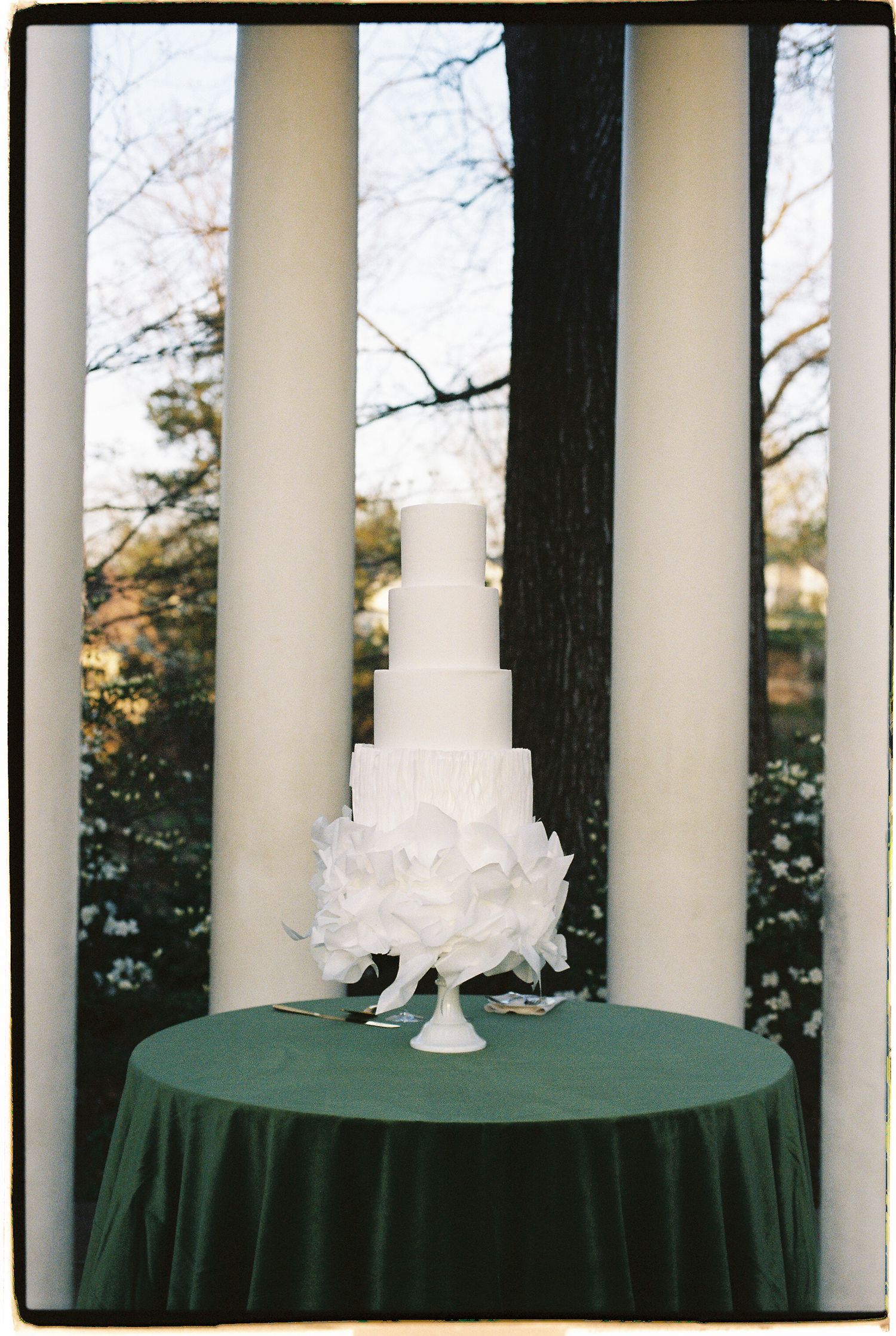 Four-tier white wedding cake with petal details on green velvet table between classic white columns.