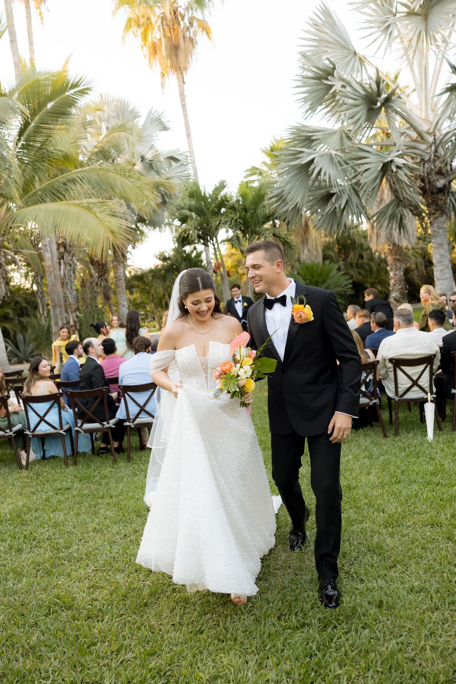 Newlyweds walk down grassy aisle after outdoor tropical wedding ceremony at golden hour.