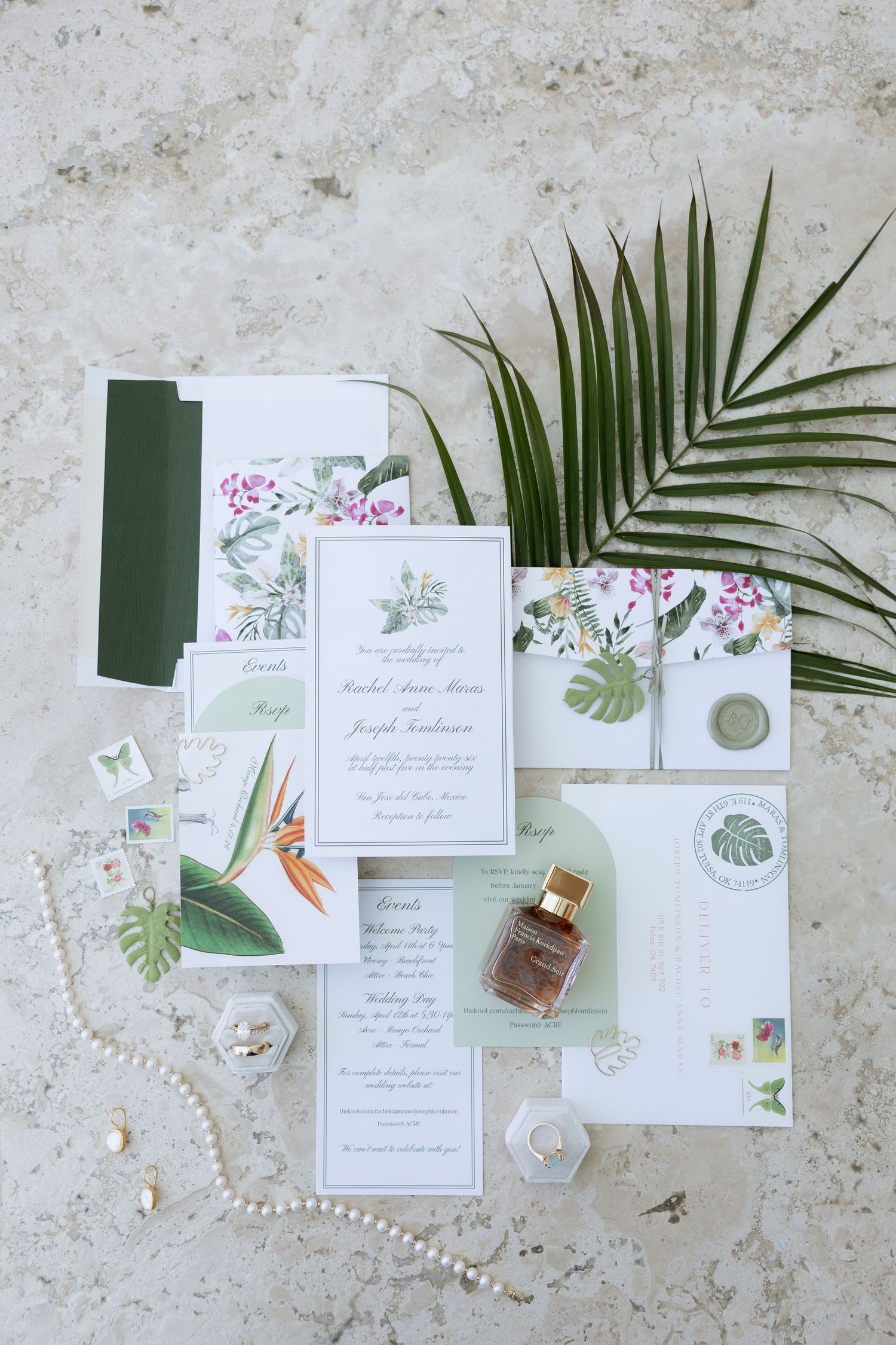 Tropical wedding invitation suite with floral designs, palm leaf, pearls, and perfume bottle on marble surface.
