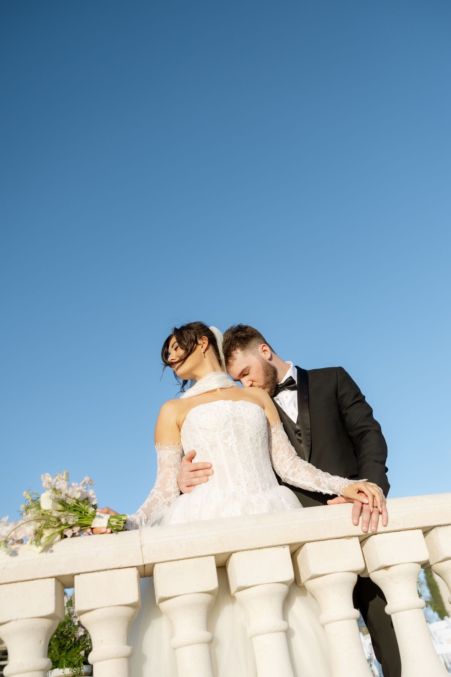 Bride and groom embrace on a white balustrade terrace under a clear blue sky on their wedding day.
