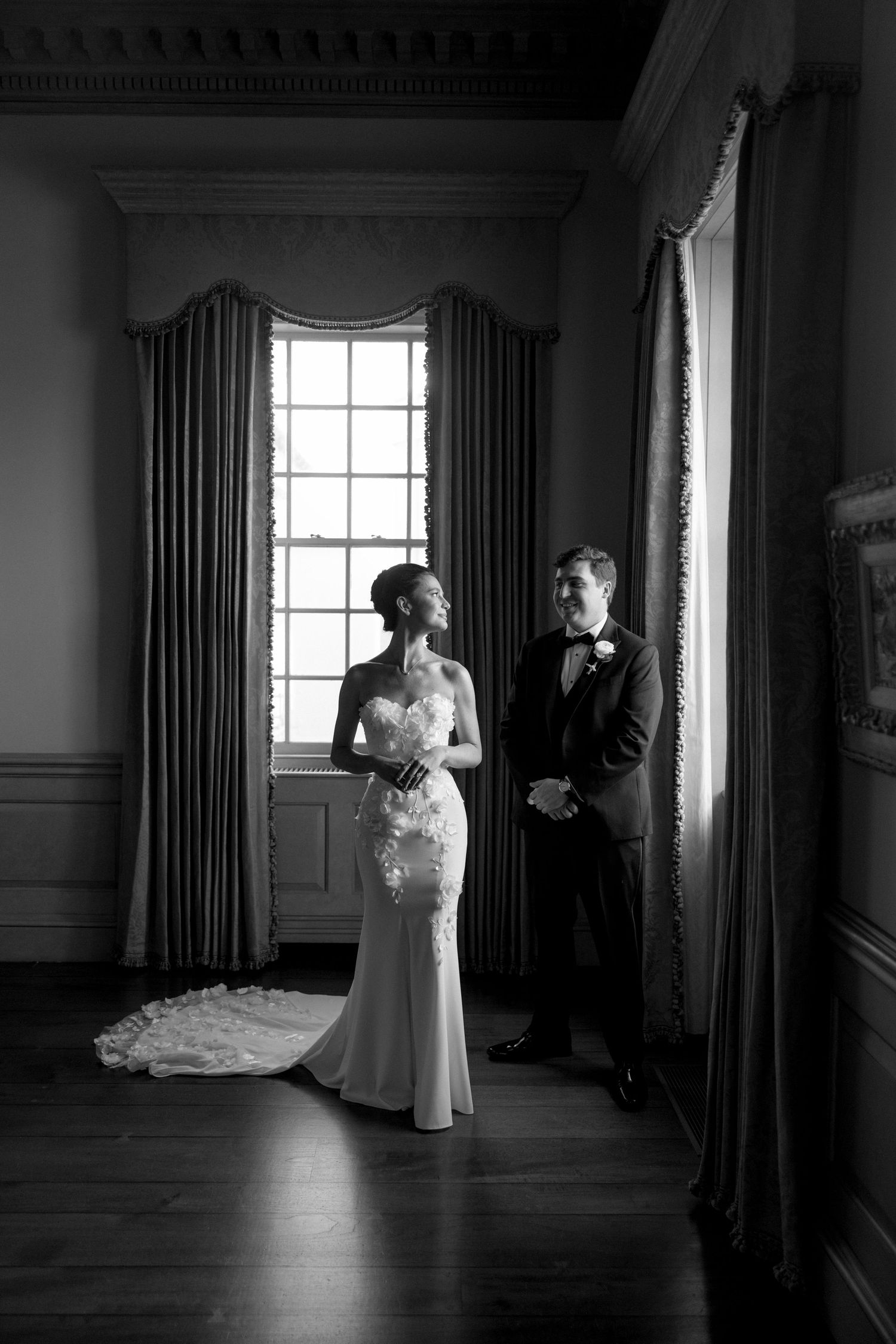 Elegant black and white portrait of bride and groom standing by tall windows in historic venue.