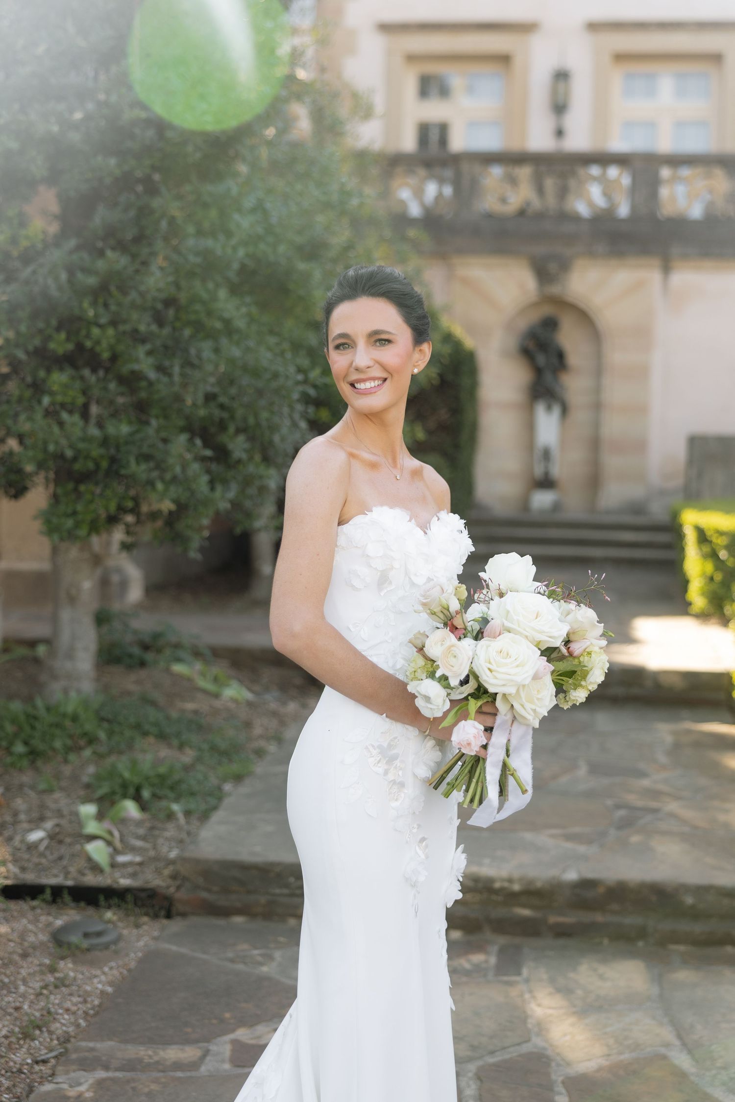 Smiling bride in a strapless white gown holds a white floral bouquet at an outdoor venue.