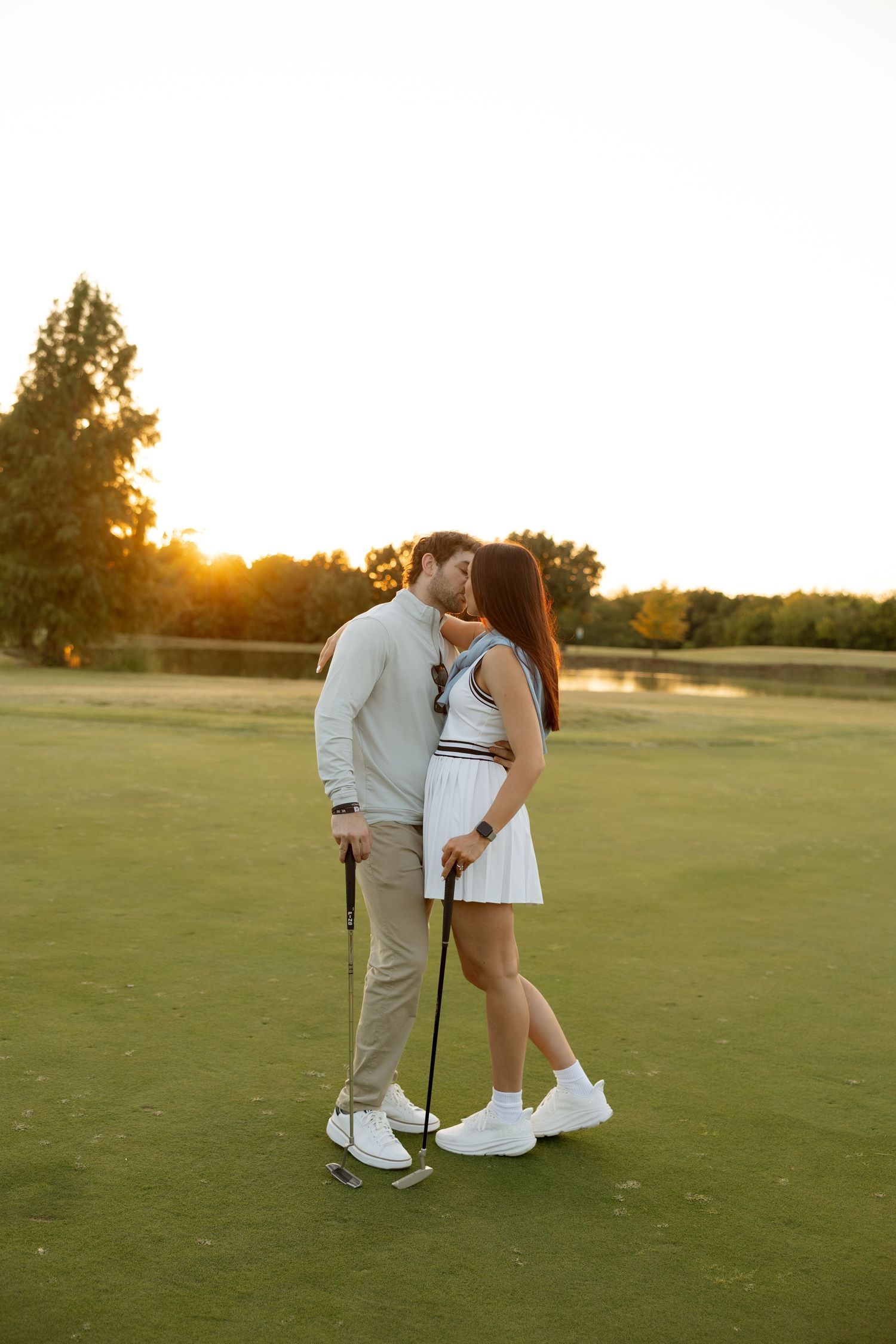 Couple in coordinated white outfits embracing on a golf course at sunset.