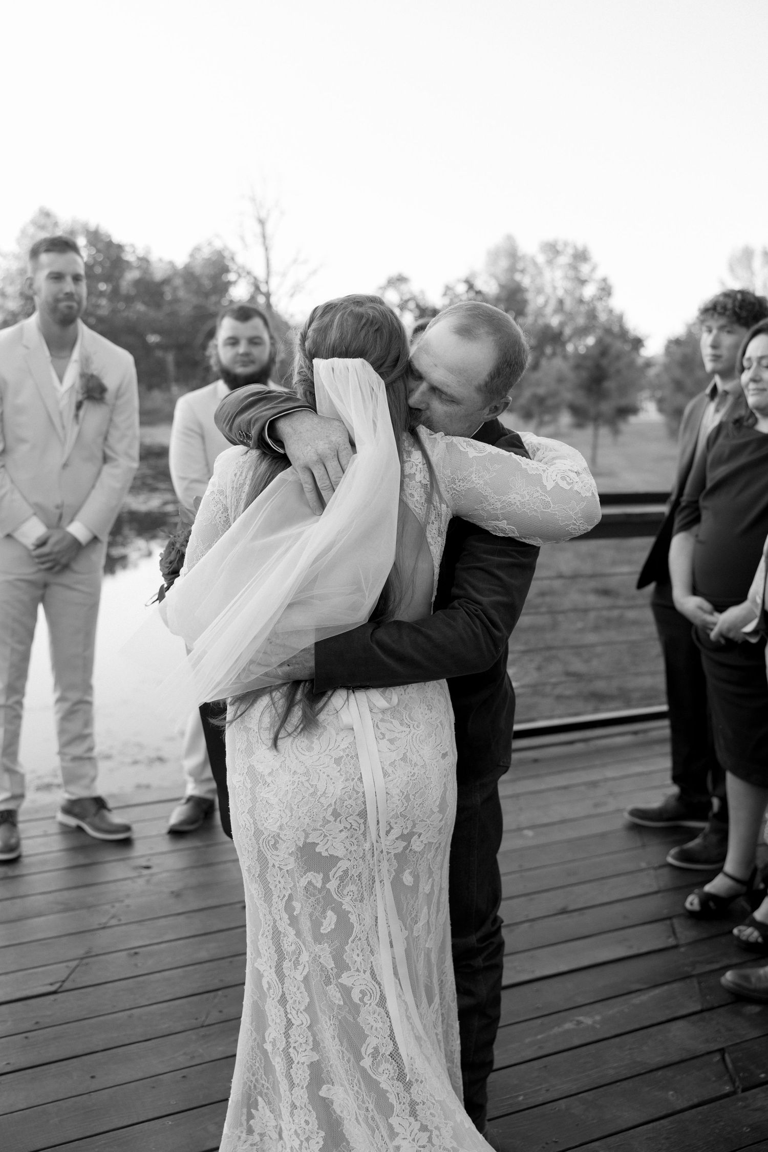 A couple shares an emotional embrace during their wedding ceremony on a wooden dock.