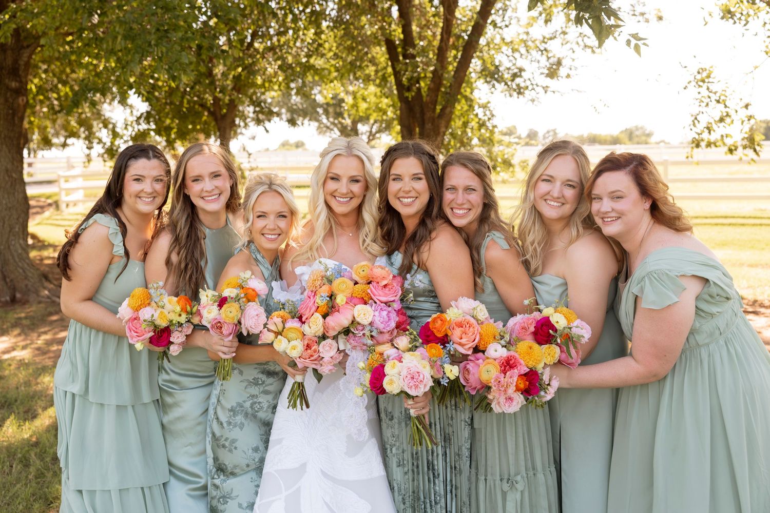 Wedding party wearing sage green bridesmaid dresses pose together with colorful bouquets at sunset.
