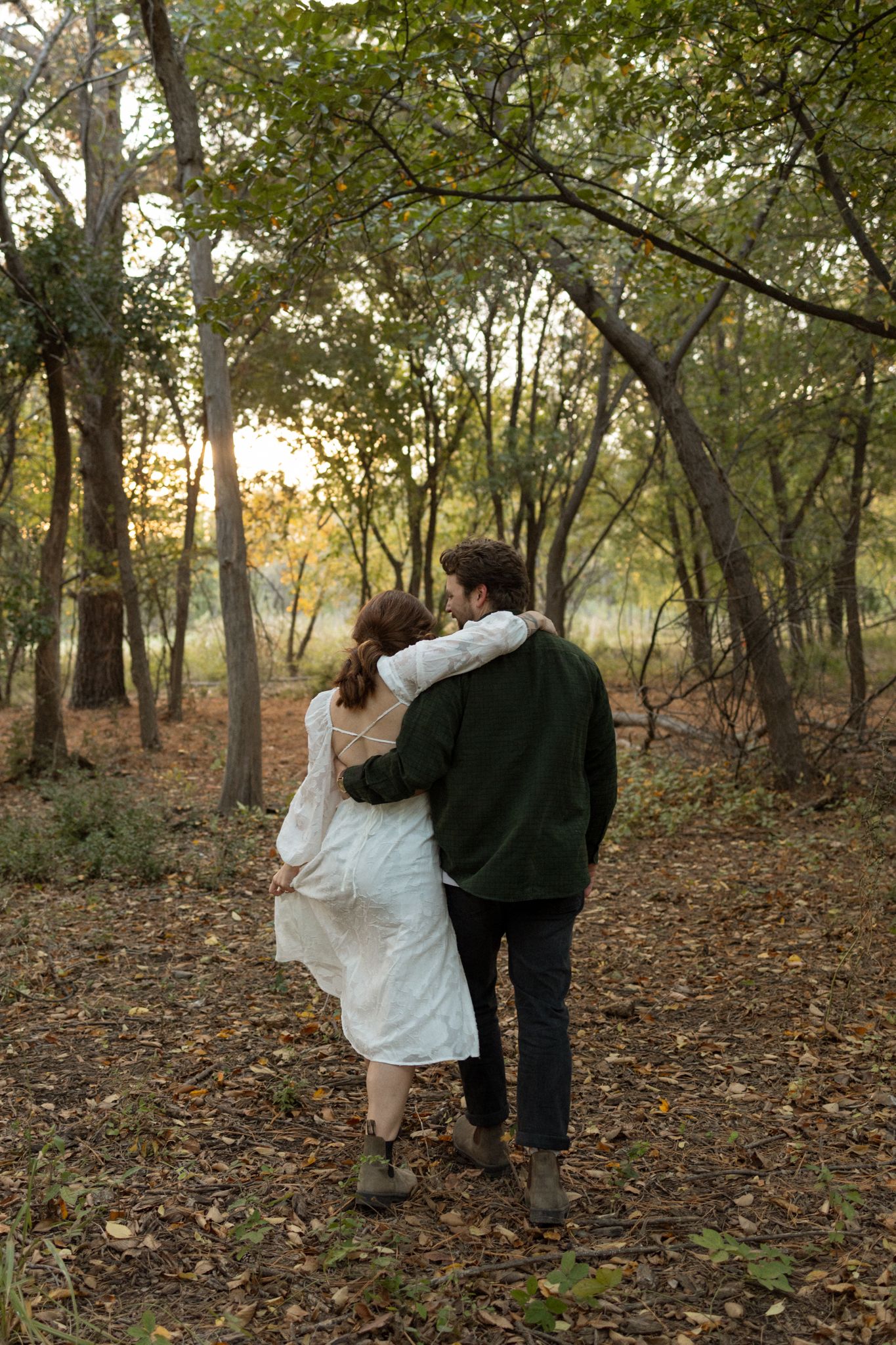 A couple embraces while walking through a wooded path during golden hour.