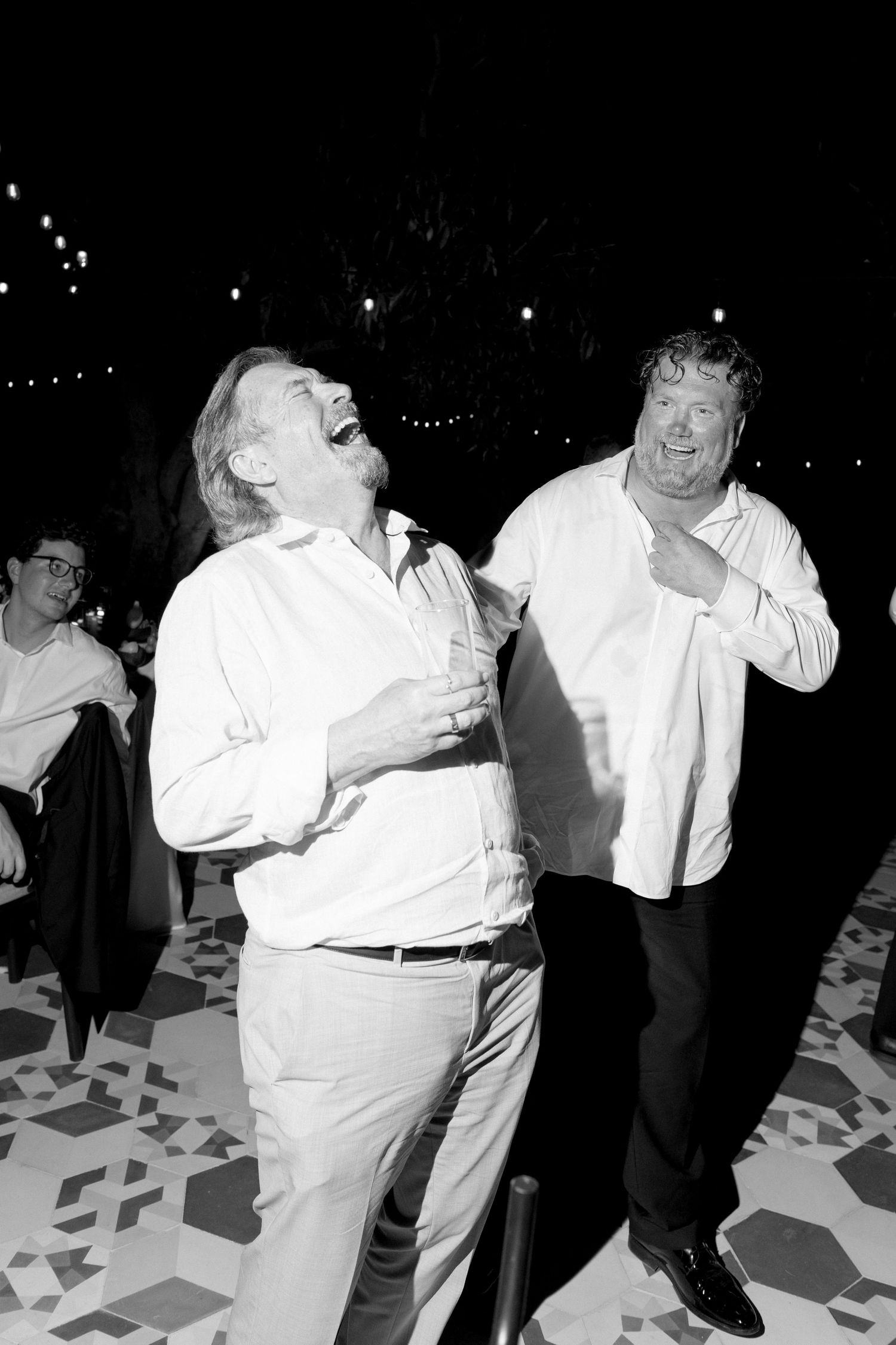 Two men in white shirts laugh and dance joyfully at an outdoor evening wedding reception.
