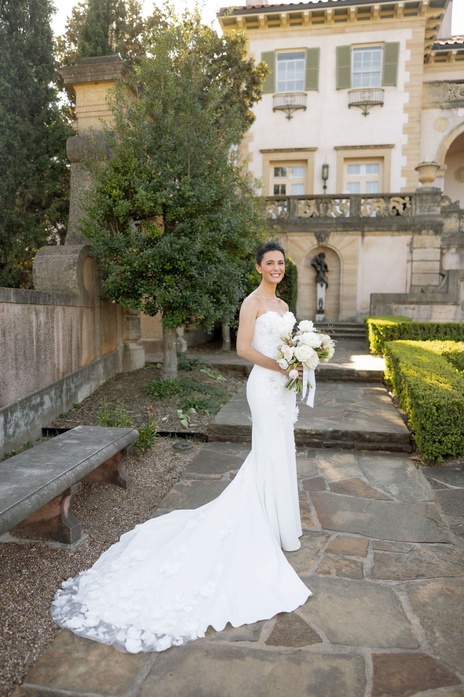 Bride in a white mermaid gown with train holds bouquet in elegant garden estate.