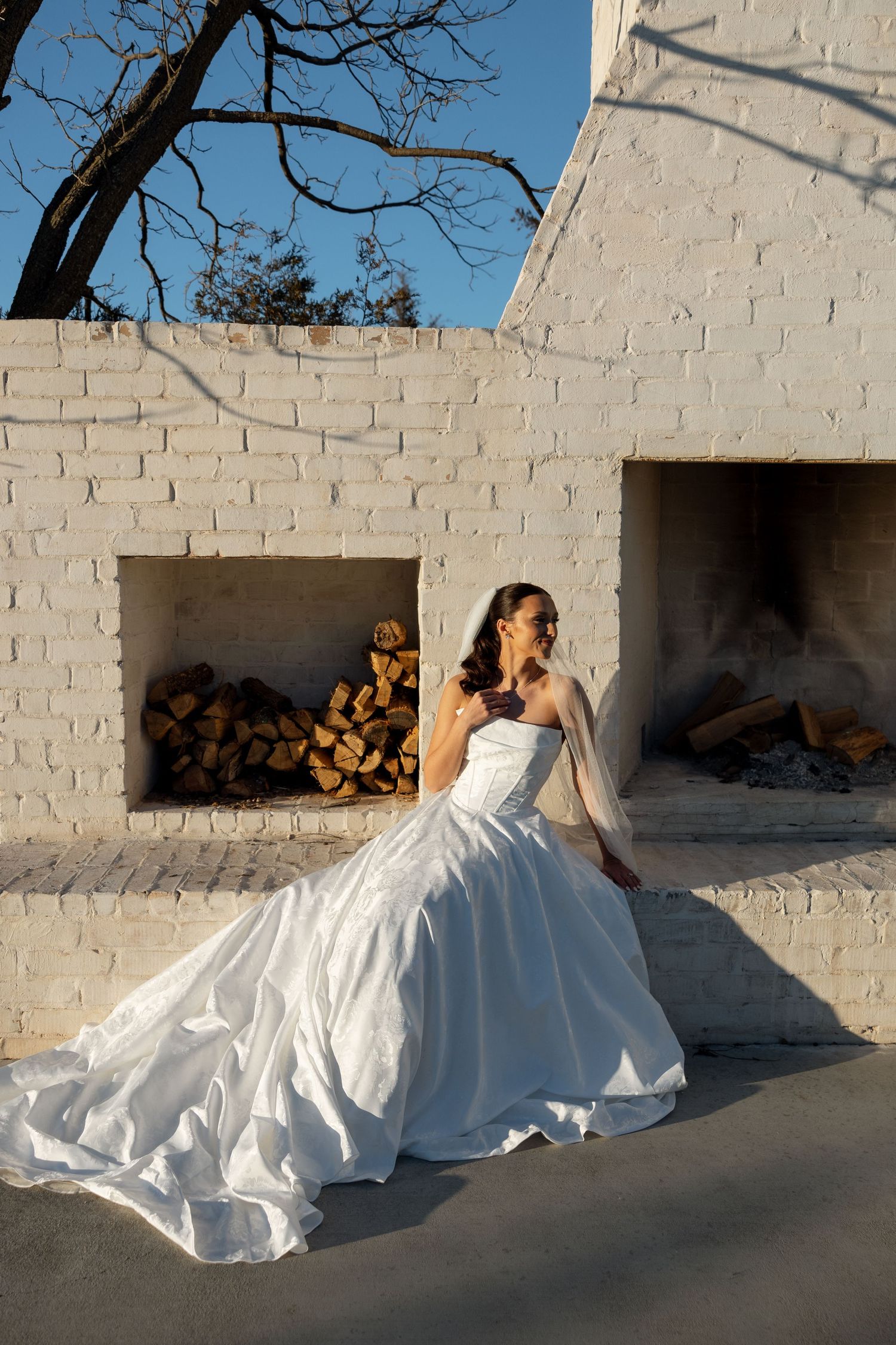 Elegant white wedding gown with dramatic train displayed against rustic white brick wall with firewood storage.