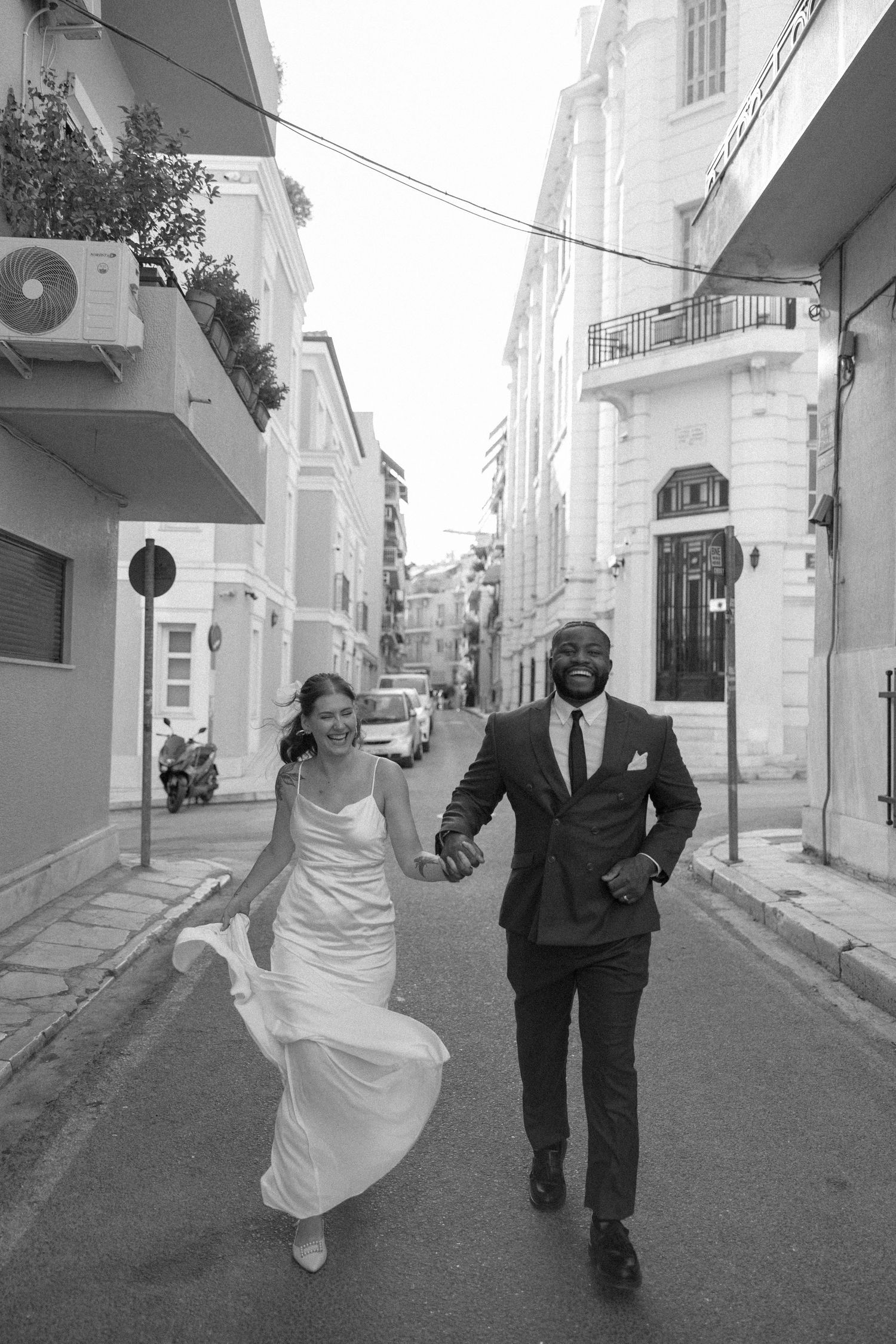 Black and white photo of couple running together through quaint city street lined with buildings.