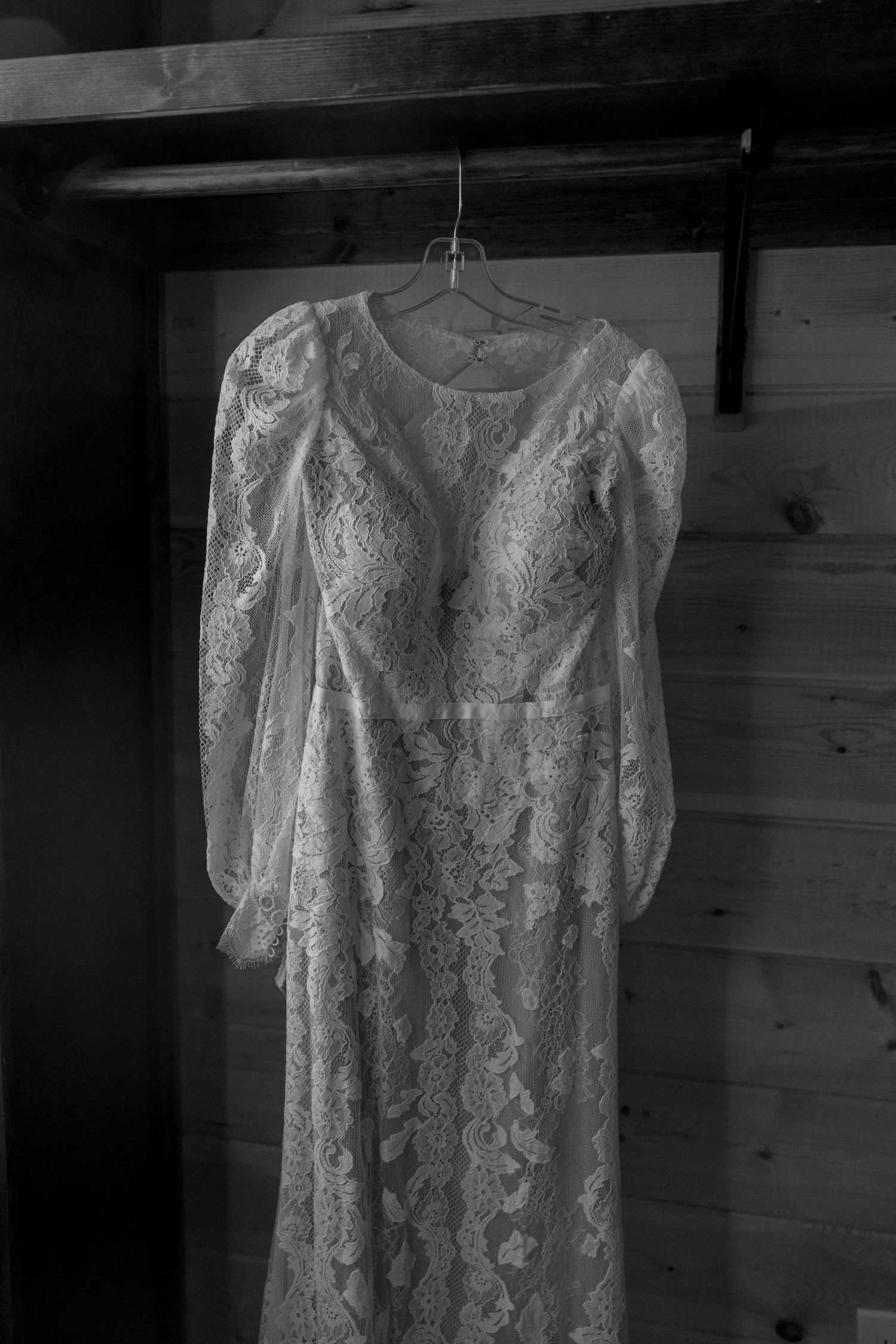 Vintage lace wedding dress hanging in a rustic wooden room, captured in black and white.