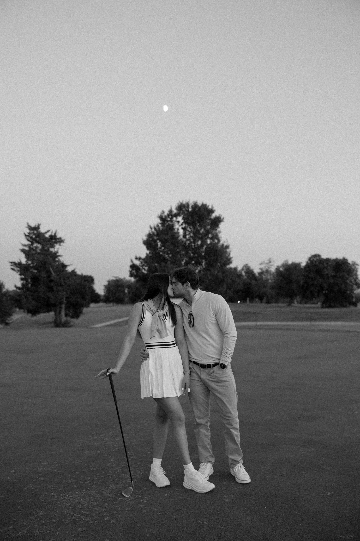 A romantic couple shares a tender moment on a golf course at dusk with the moon visible in the background.