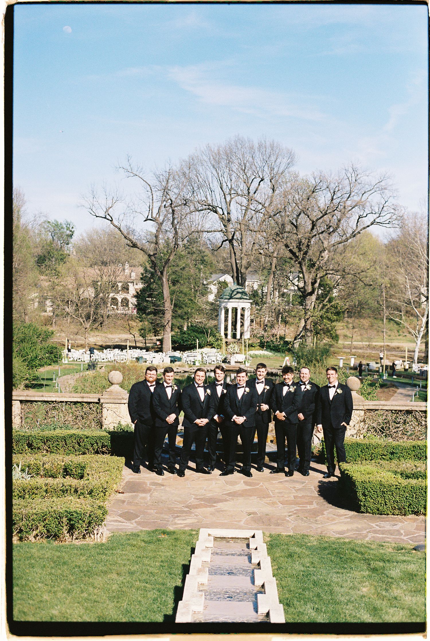 Groomsmen in black tuxedos pose in a formal garden with a gazebo and trees at an outdoor wedding.