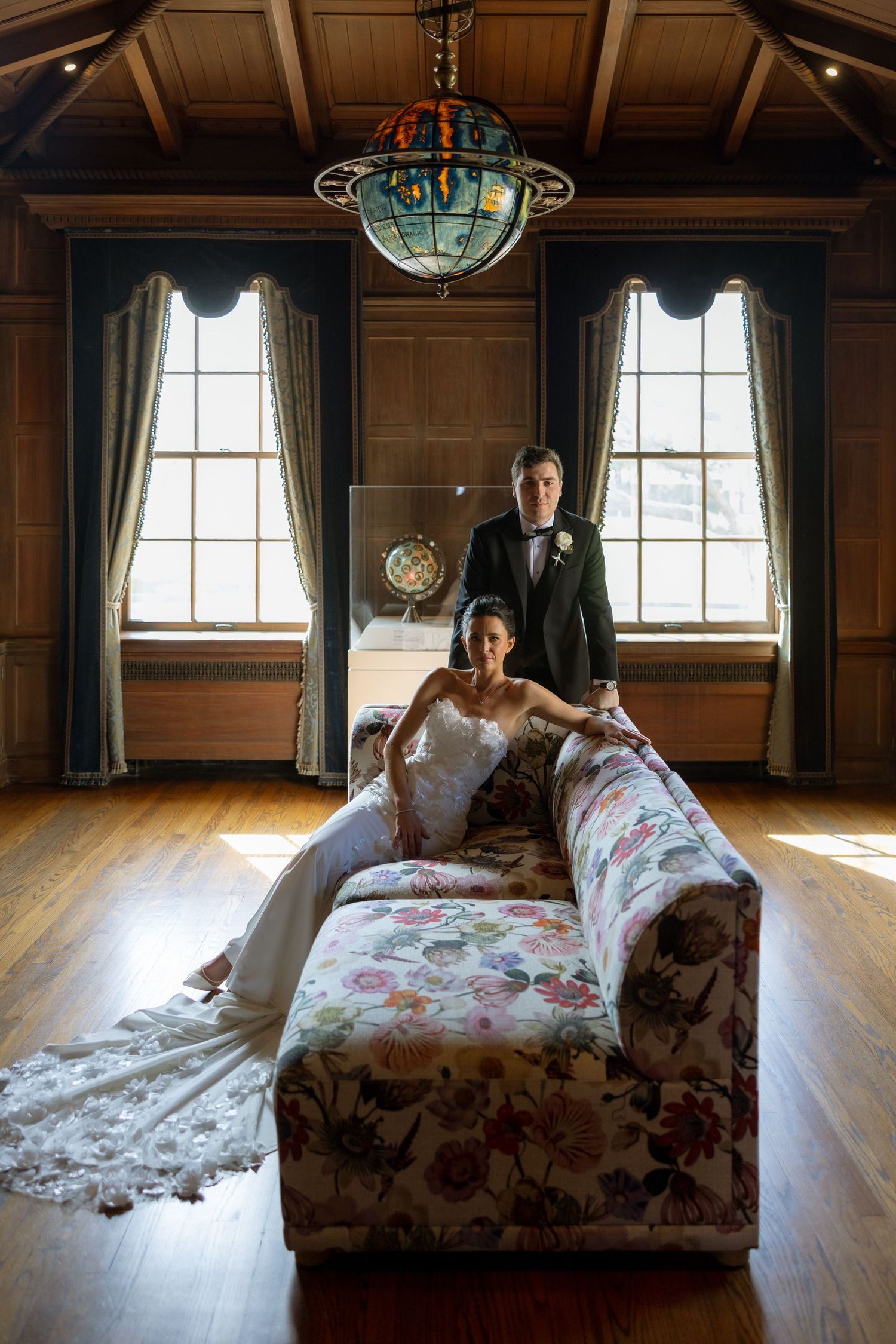 Bride reclines on floral sofa as groom stands behind her in wood-paneled room with chandelier.