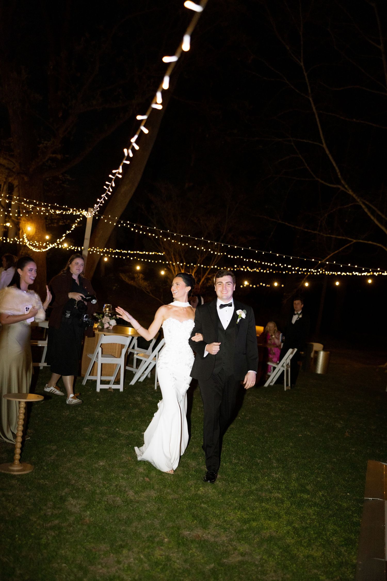 Newlyweds make grand entrance under string lights at elegant outdoor nighttime wedding reception.