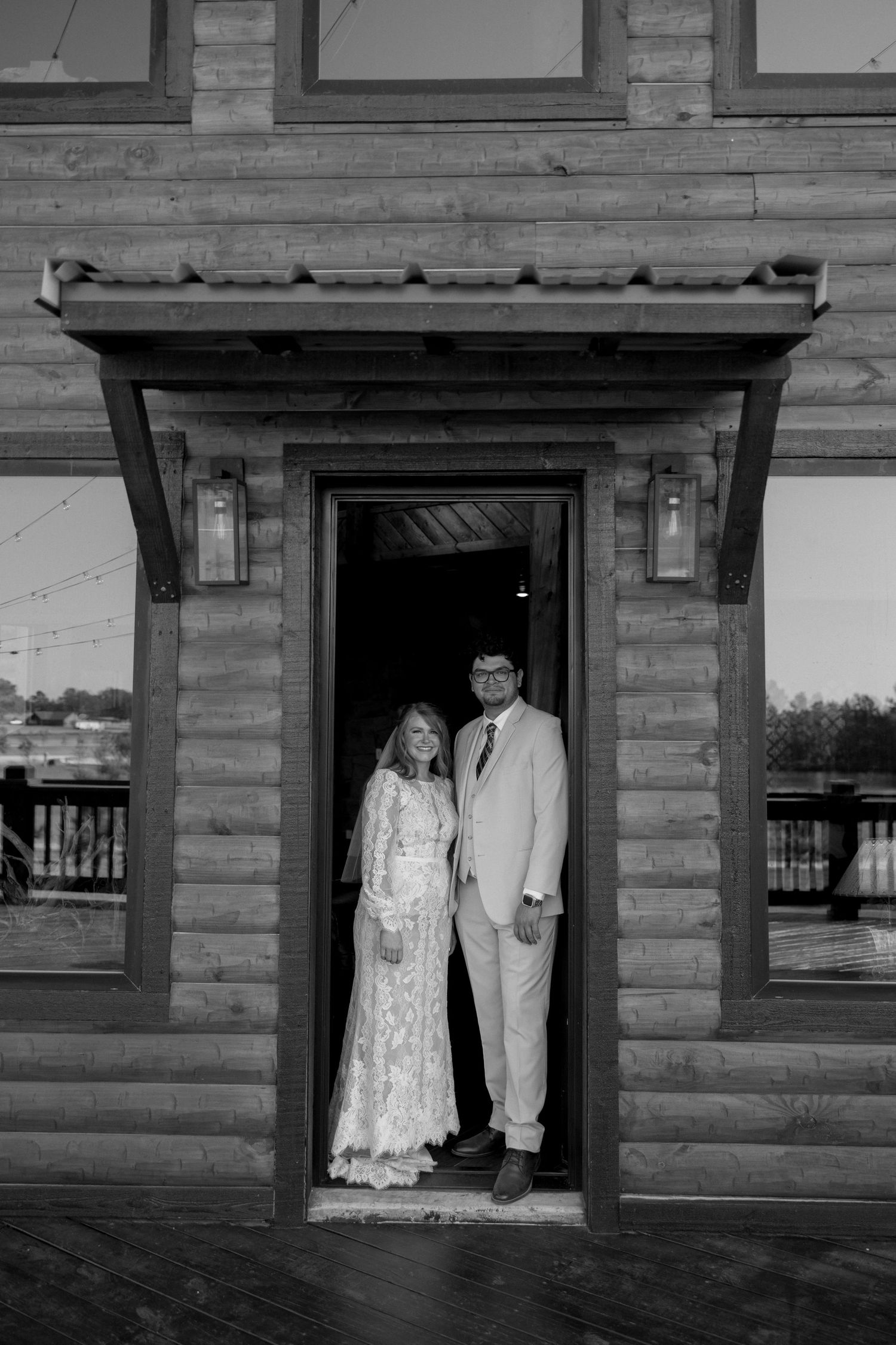 Black and white portrait of a couple standing in a rustic wooden doorframe.