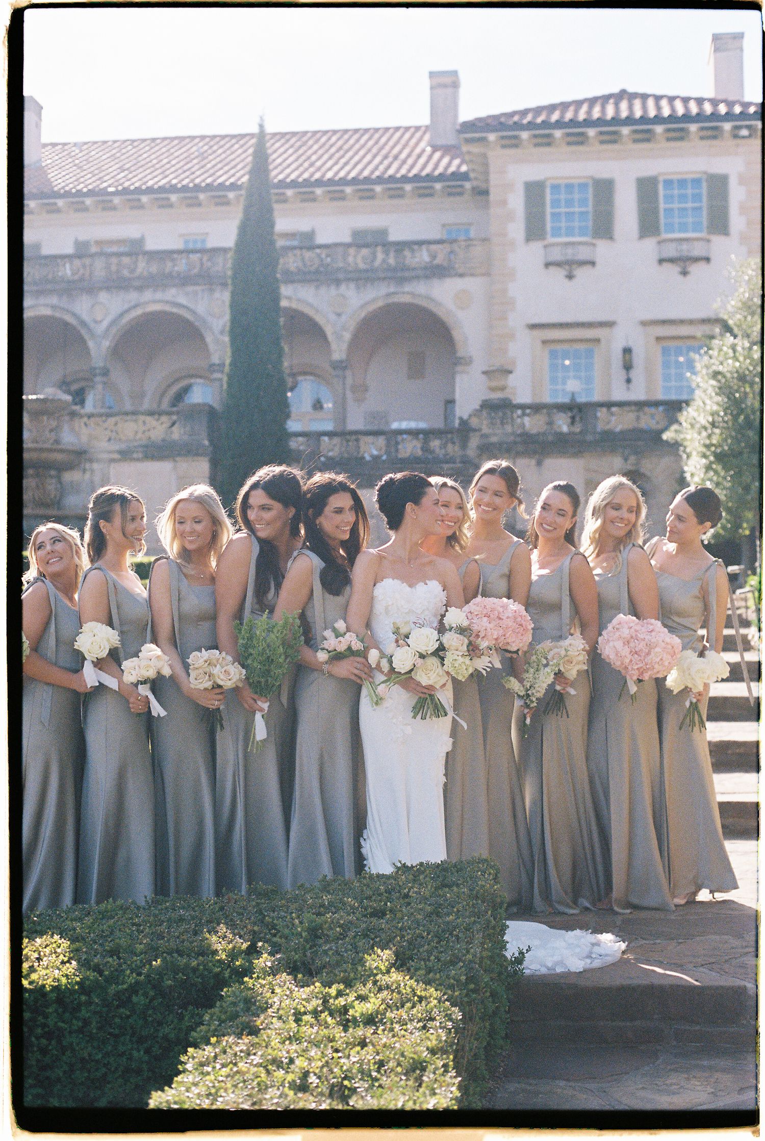 Bride poses with bridesmaids in sage green gowns holding pink and white bouquets in front of elegant venue.