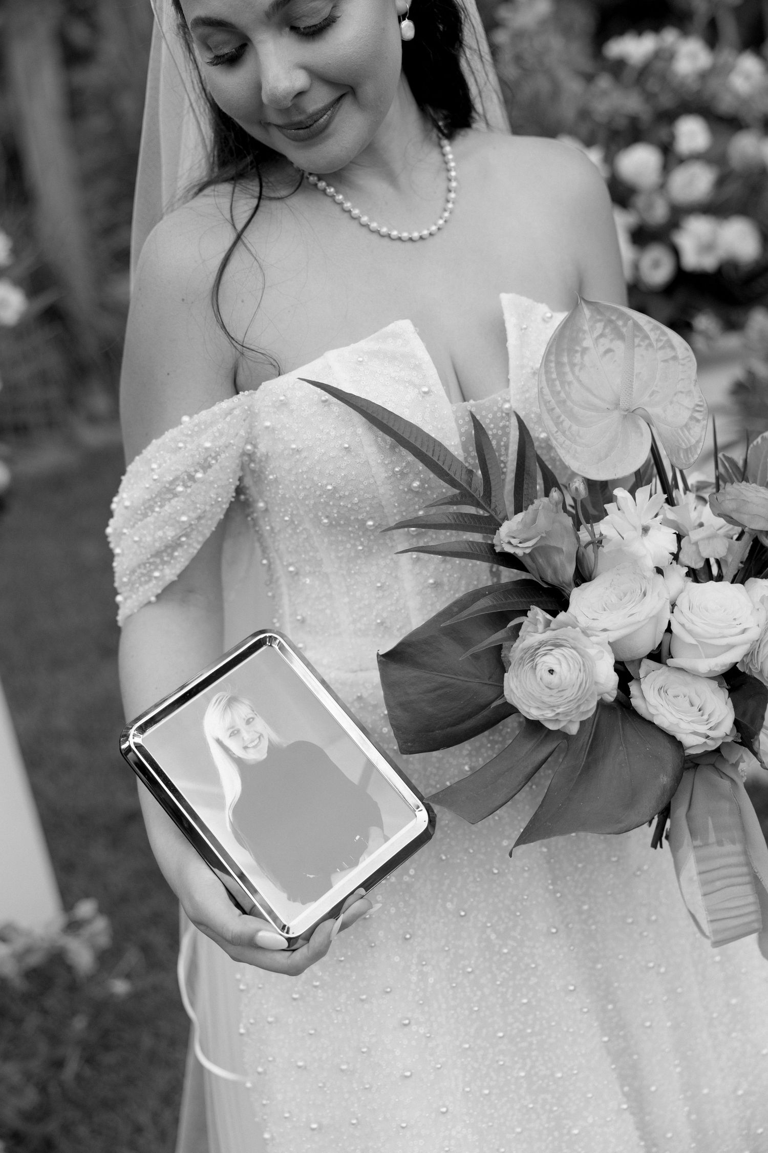 Bride in pearl wedding gown holds bouquet and framed photo of loved one at outdoor wedding ceremony.