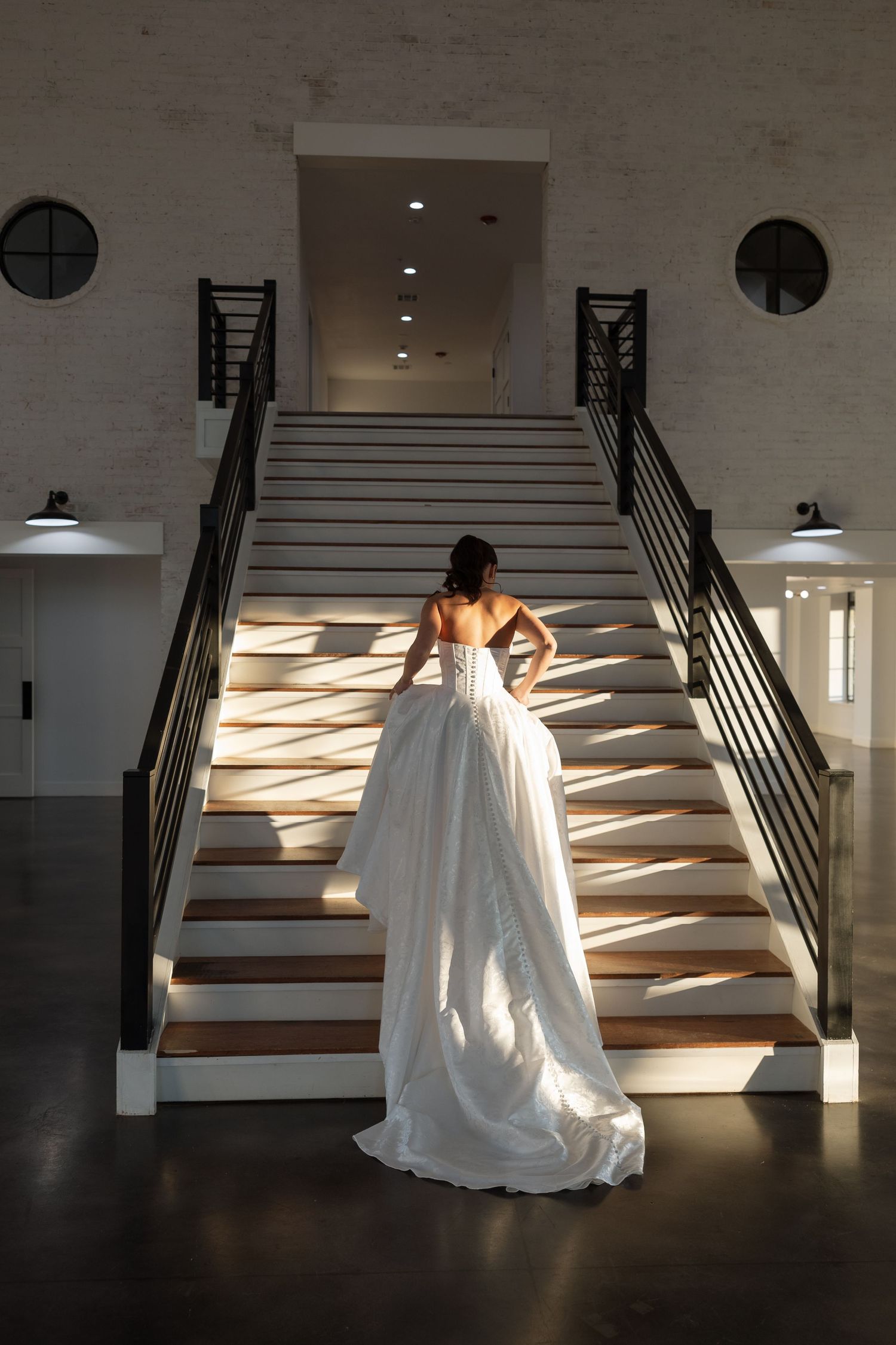 A bride in a flowing white wedding gown ascends a dramatically lit staircase with circular windows.