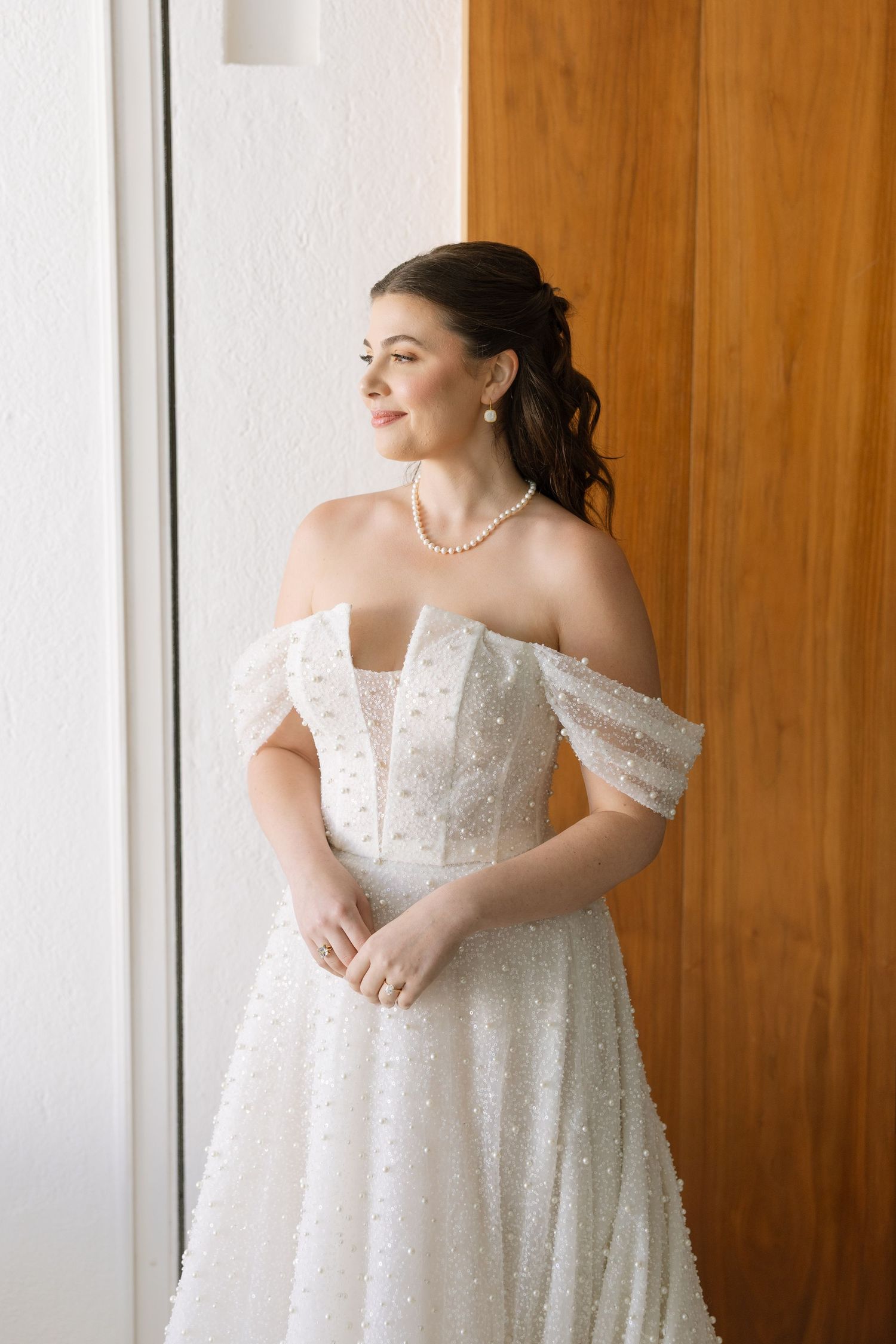 Elegant bride in off-shoulder pearl-embellished wedding gown wearing pearl necklace gazes out window in soft natural light.