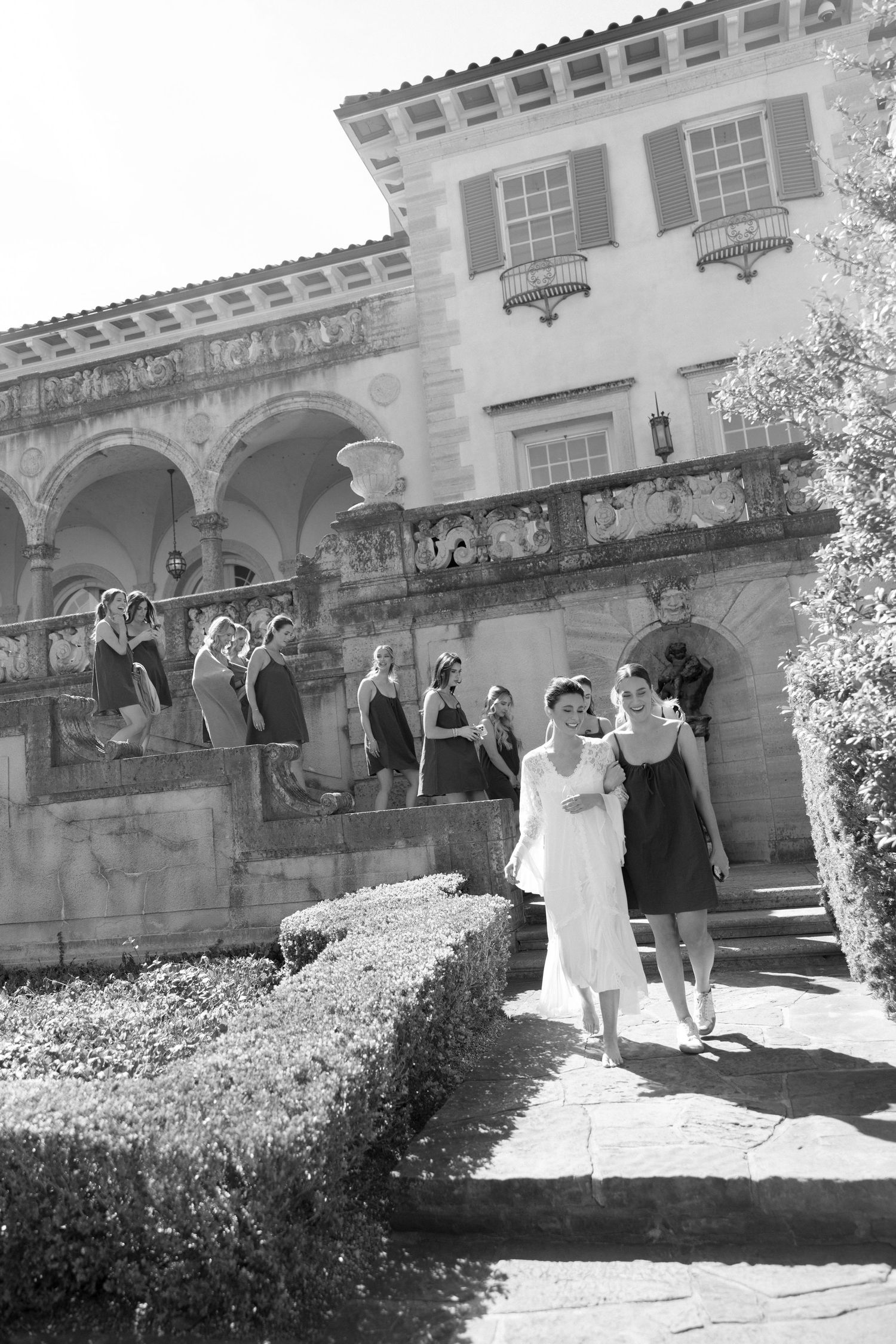 Black and white photo of a bride and bridesmaids descending steps of a grand historic mansion on a sunny day.