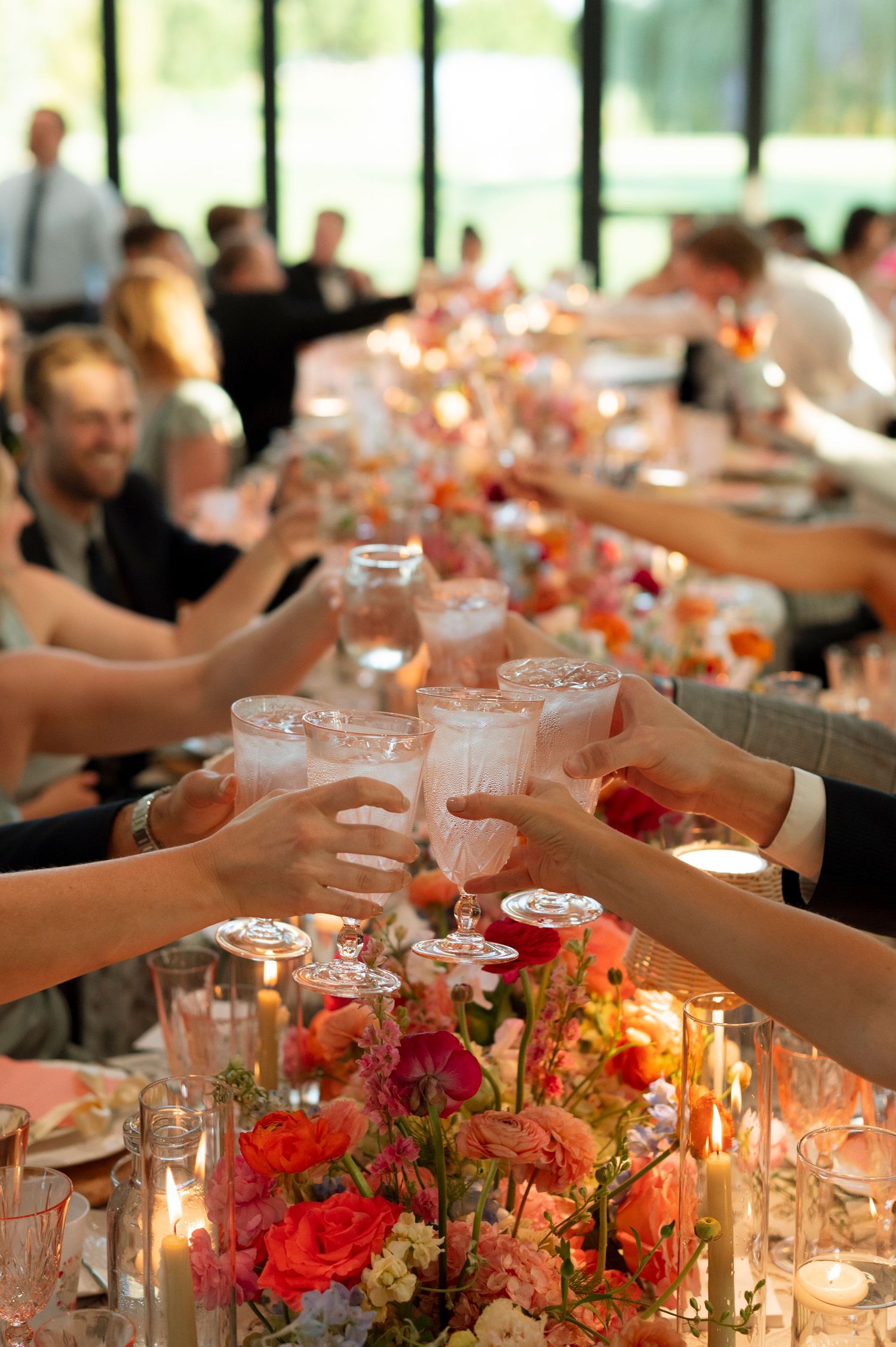 Wedding reception table with coral floral centerpieces and glowing candles as guests raise glasses in celebration.
