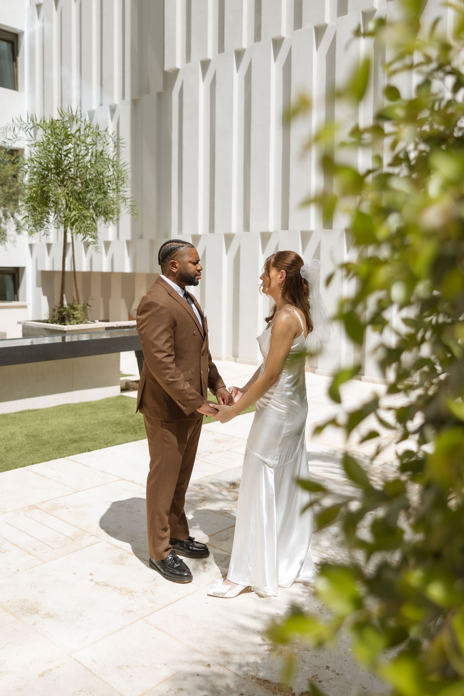 A couple holds hands while standing together by a modern white building with tropical landscaping.