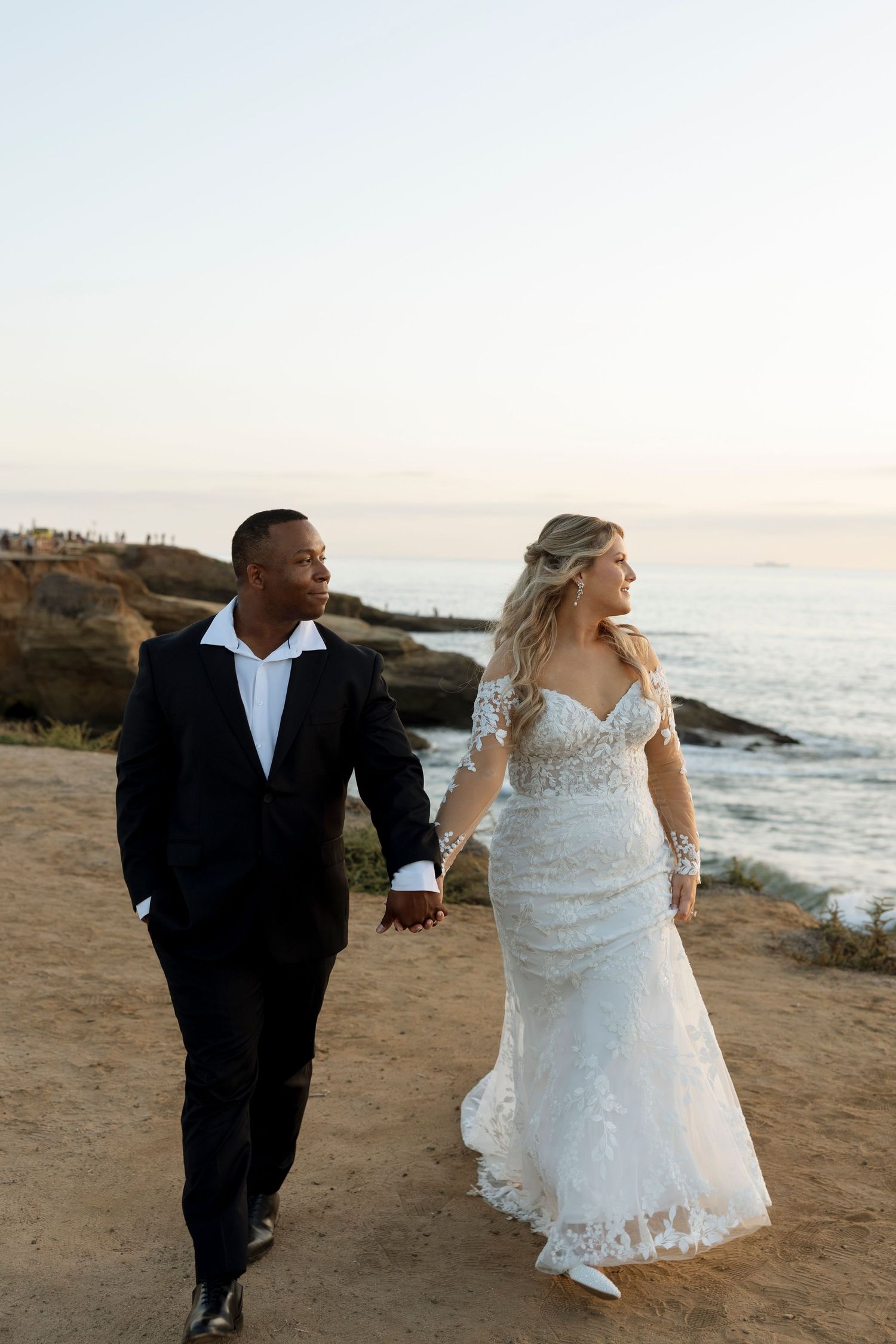 Newlyweds walking hand in hand along a scenic coastal cliff at sunset with ocean waves crashing below.