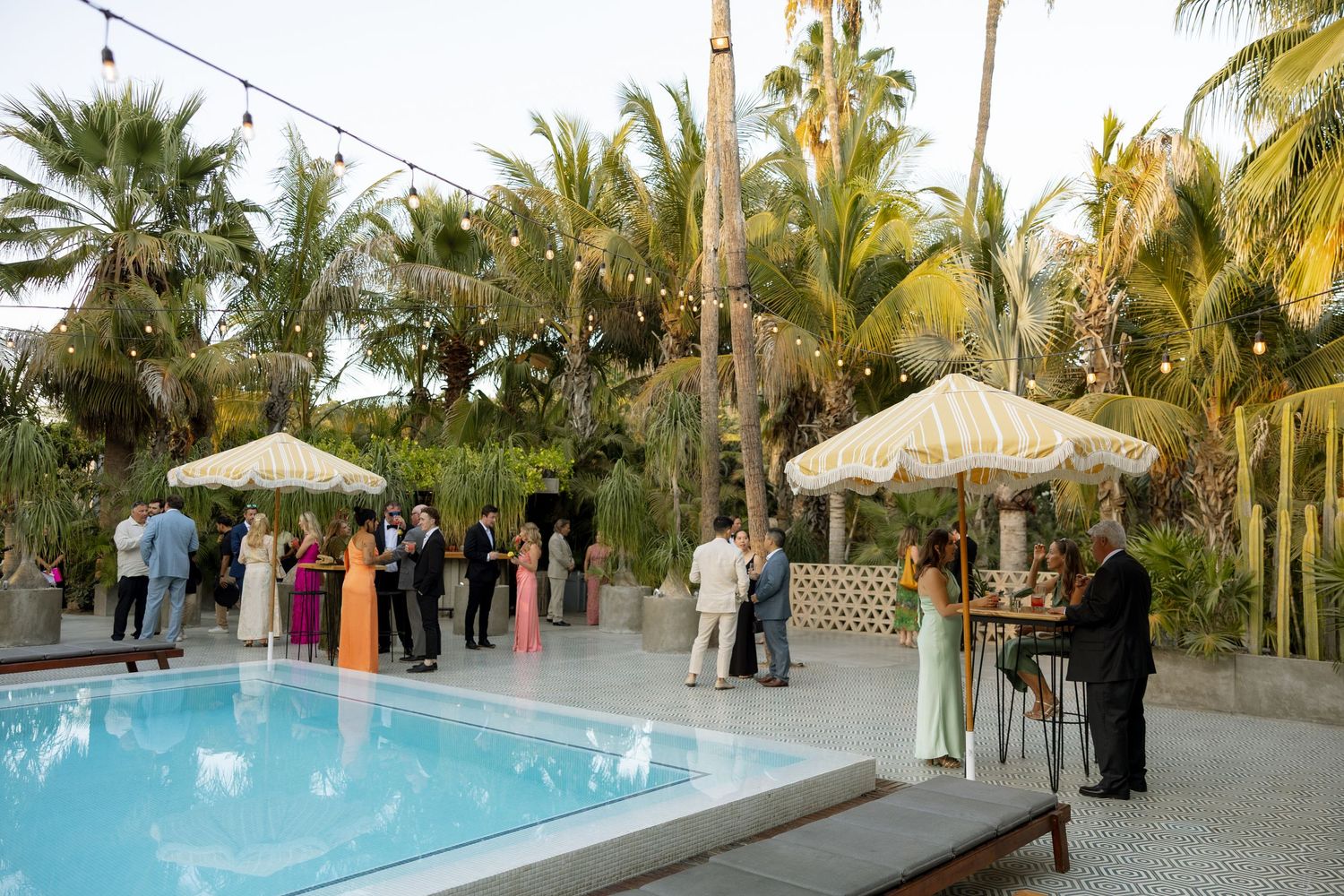 Elegant outdoor wedding reception by a pool with palm trees, string lights, and guests in formal attire.