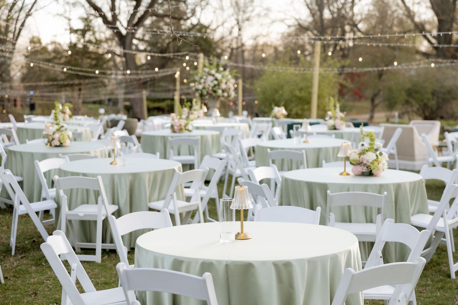 Outdoor wedding reception with sage green linen round tables, white folding chairs, and string lights.