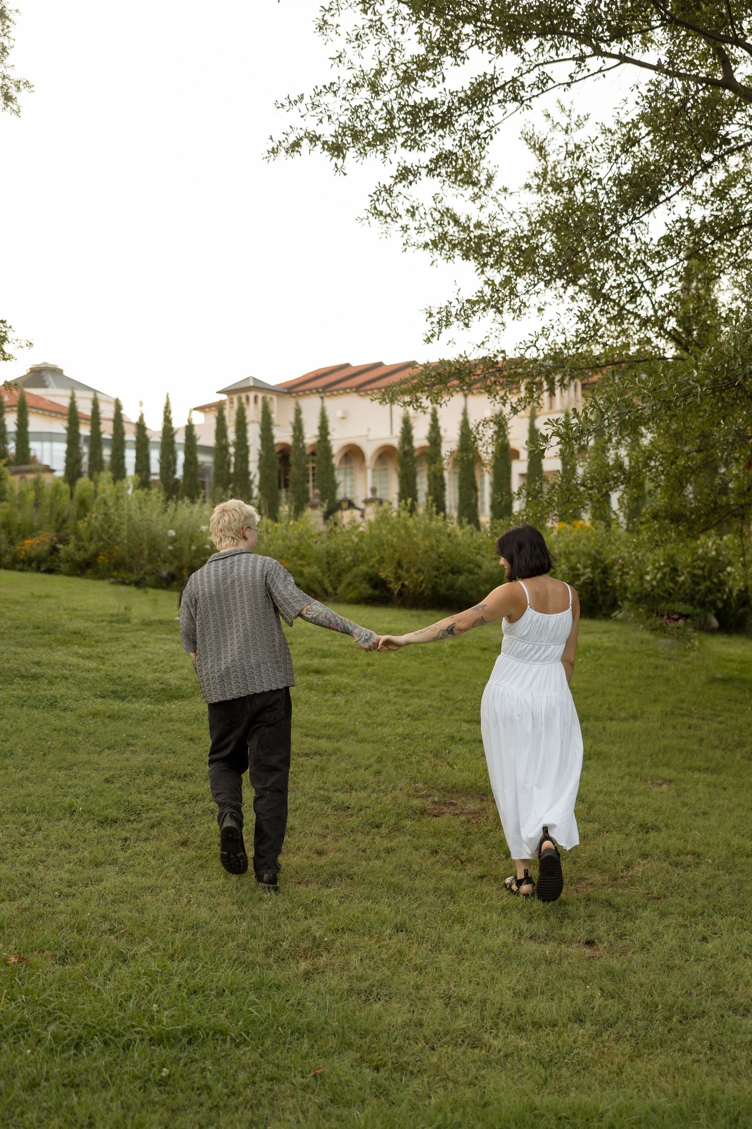 Two people walk hand in hand towards a Mediterranean-style villa with cypress trees.
