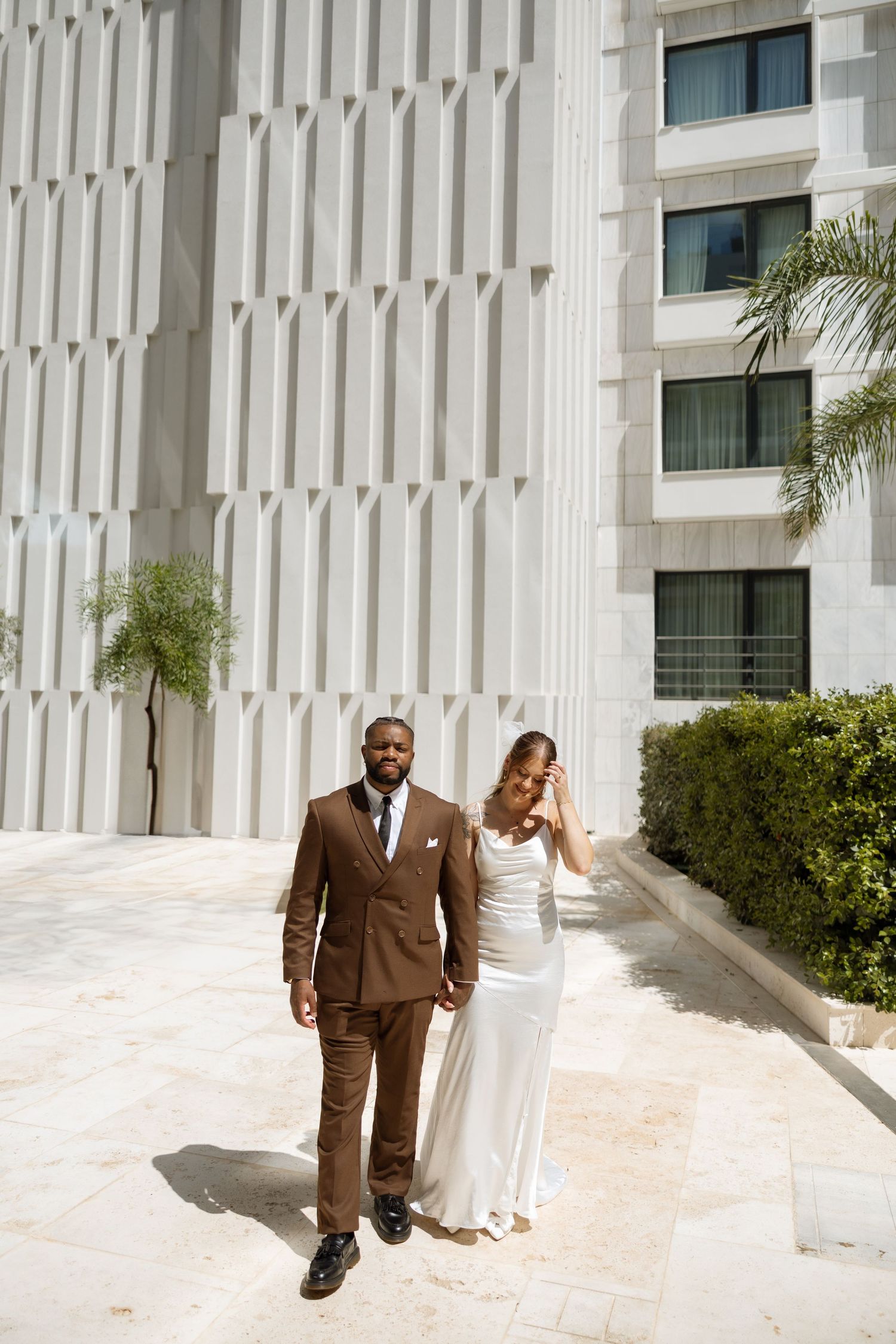 Wedding couple walking together near a modern white building with palm trees.