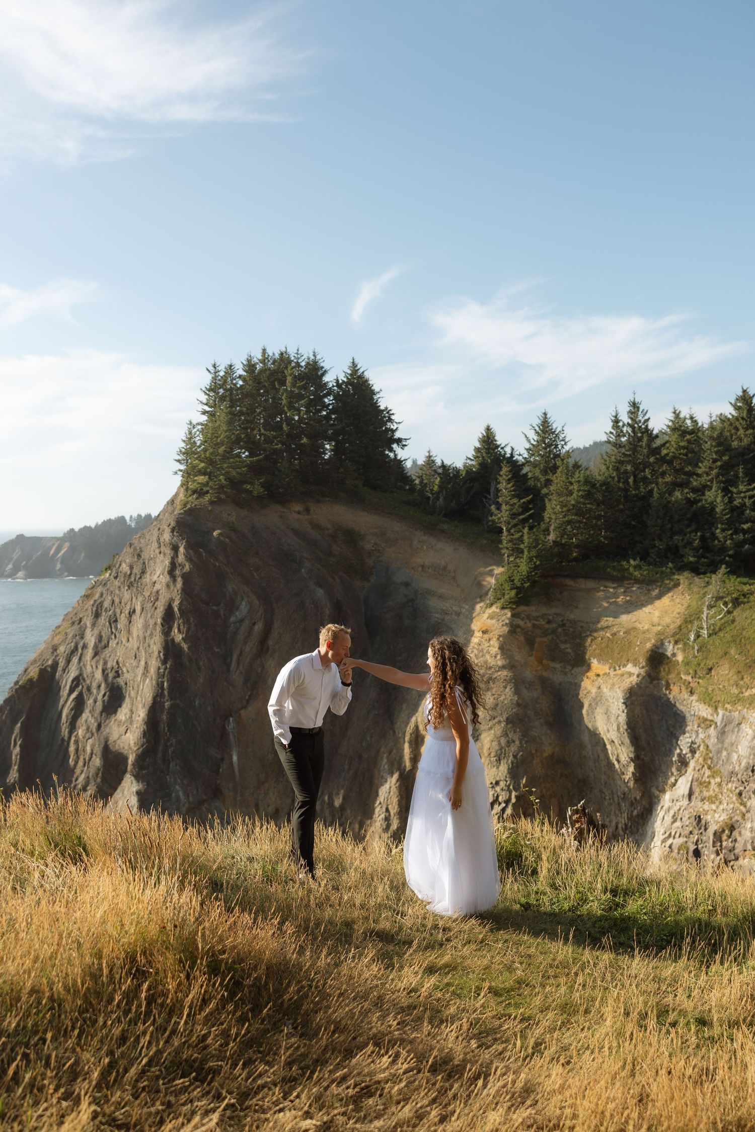 A romantic wedding couple poses together on a dramatic coastal cliff with evergreen trees and ocean views.