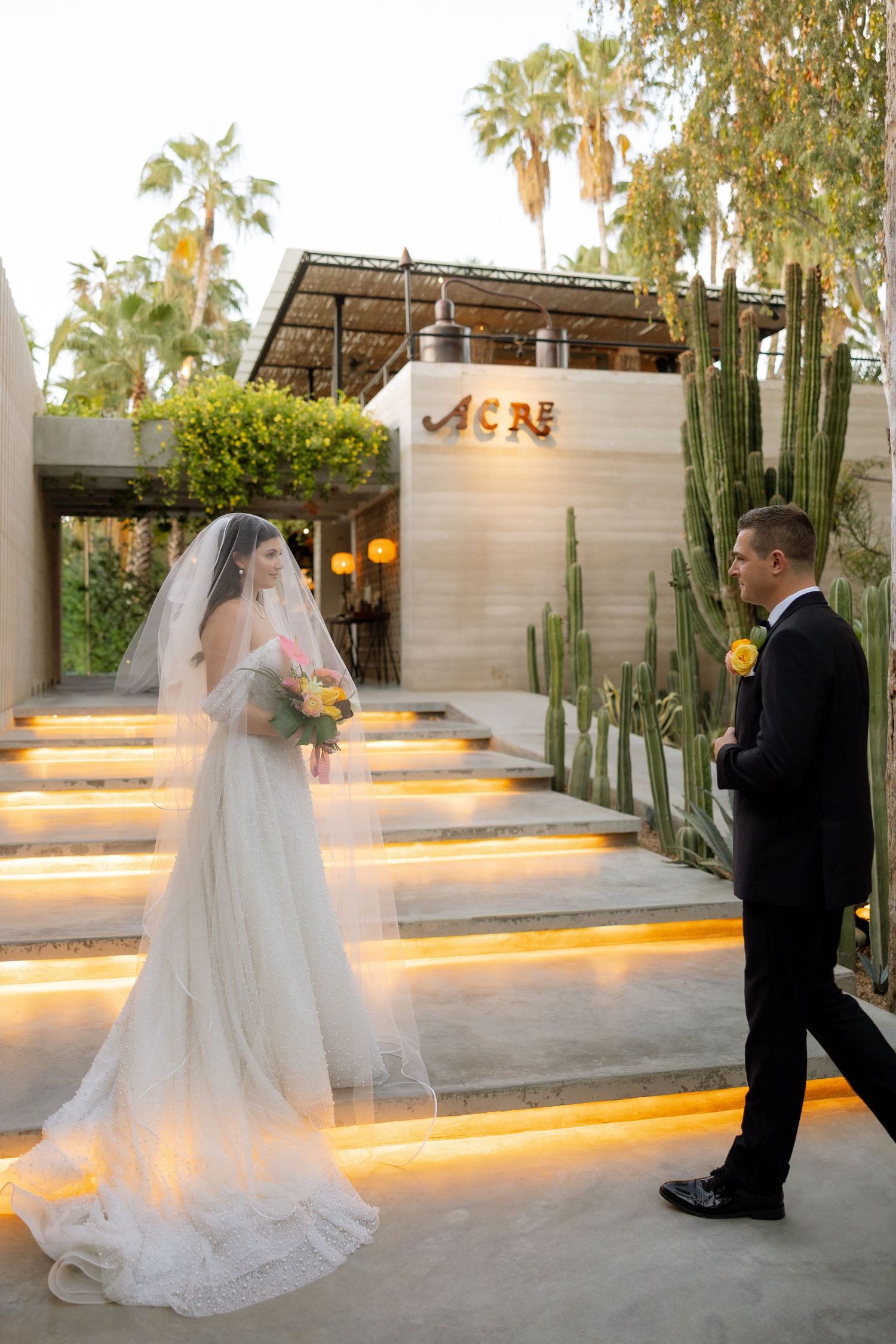 Bride in white gown and veil stands on illuminated steps at Acre venue as groom in tuxedo approaches.