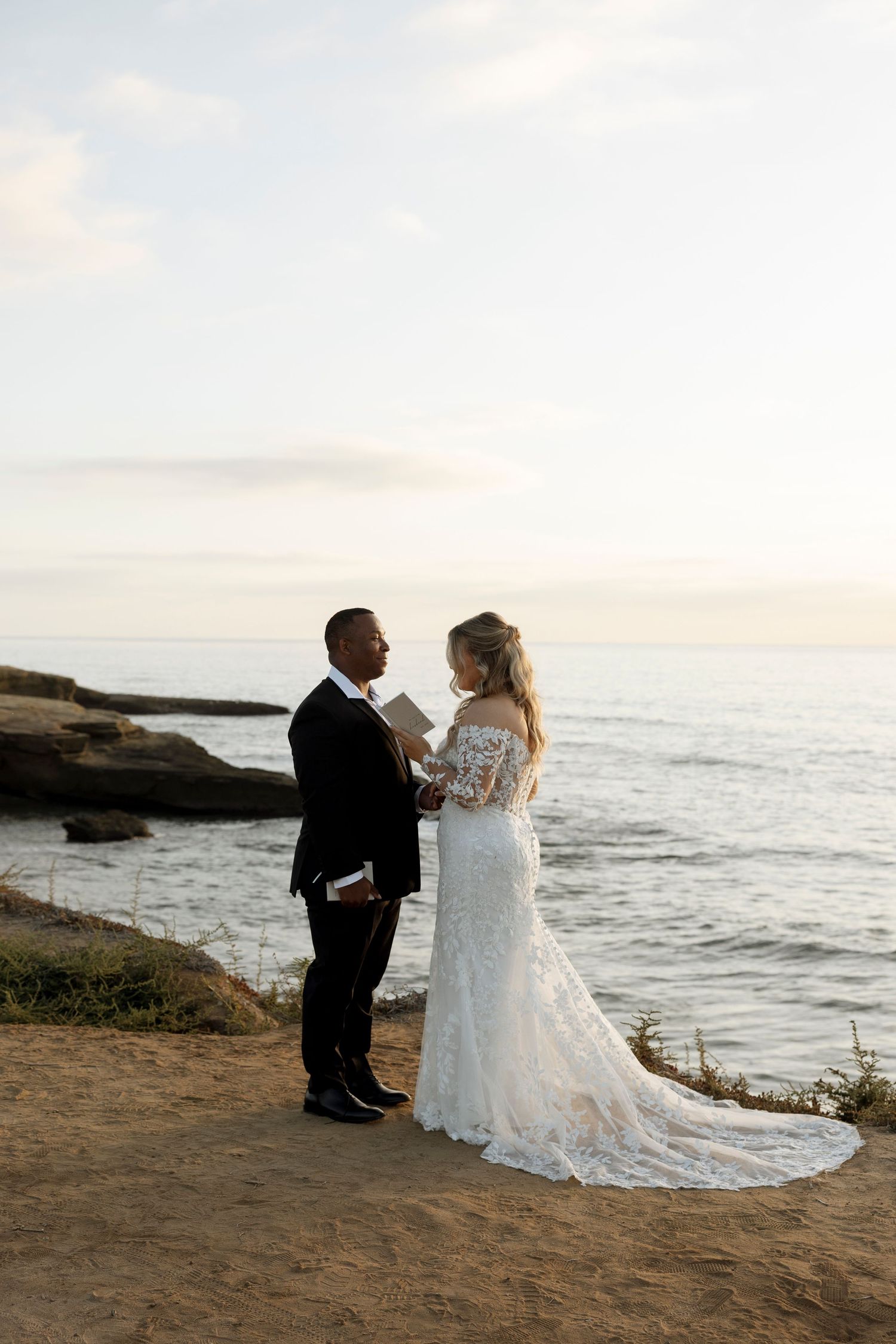 Wedding couple poses on coastal cliffs at sunset with ocean waves in background.