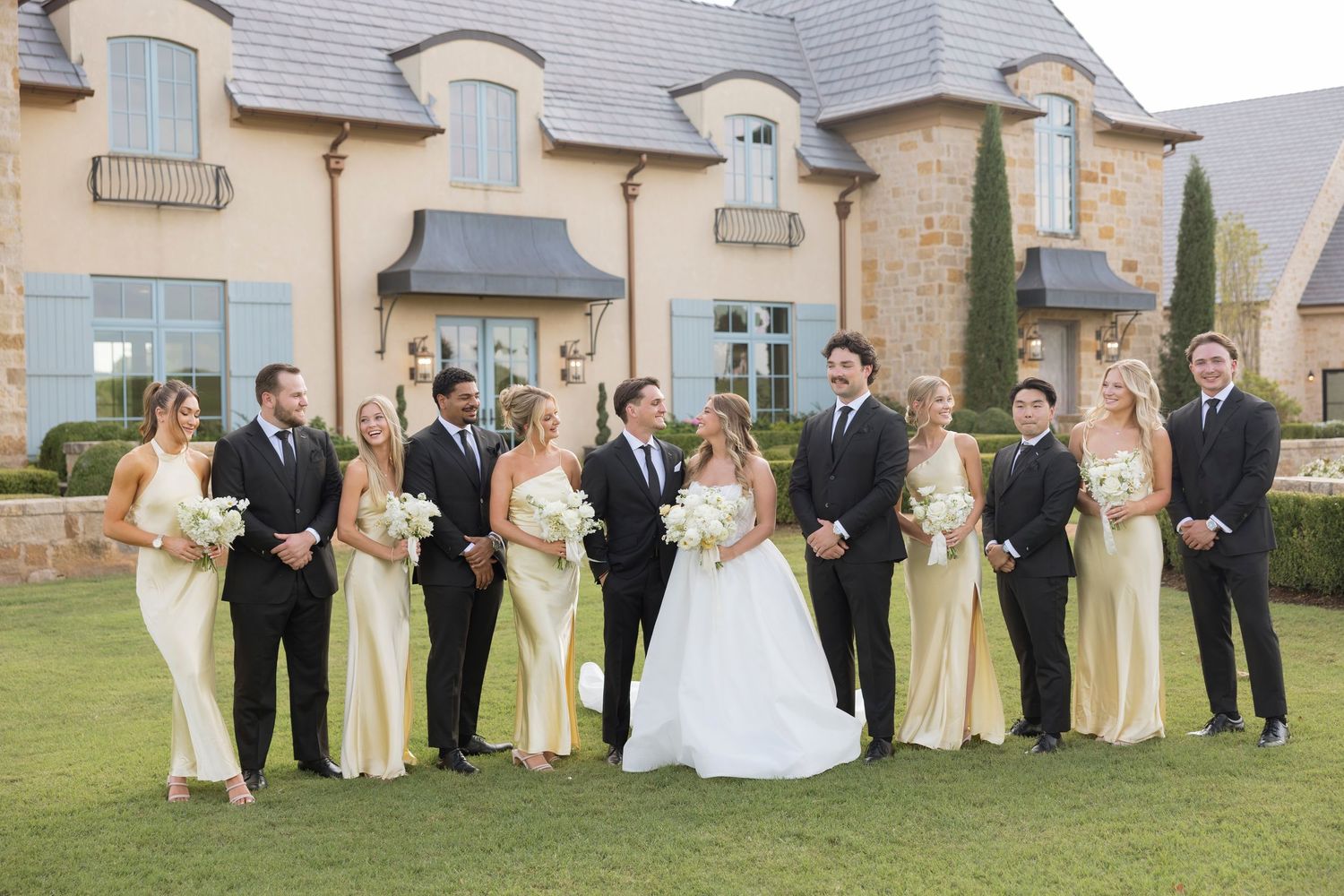 Wedding party in formal attire poses together on green lawn outside elegant stone venue during outdoor celebration.