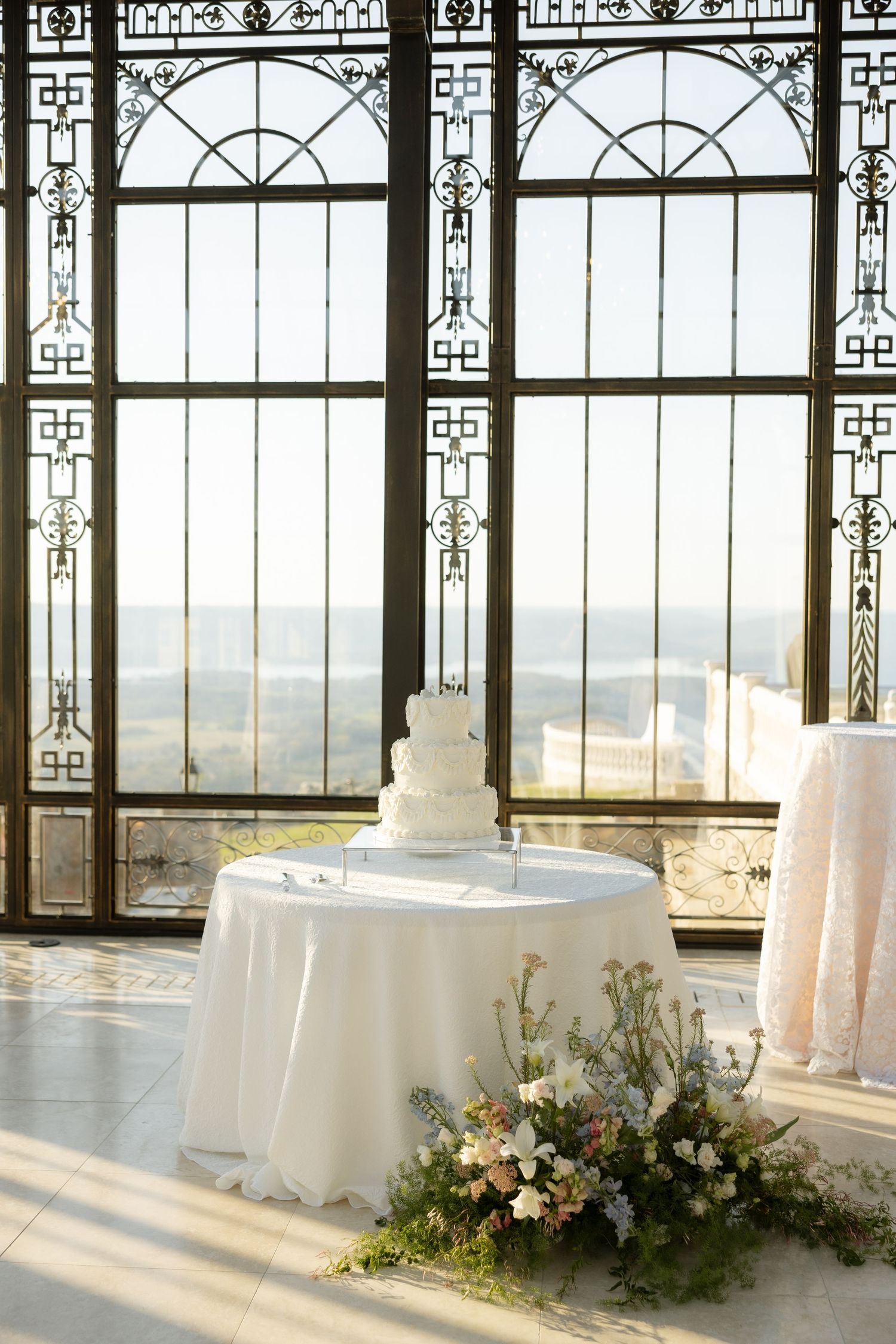 Elegant white tiered wedding cake on draped table with floral arrangement, set before ornate iron-framed windows with scenic view.