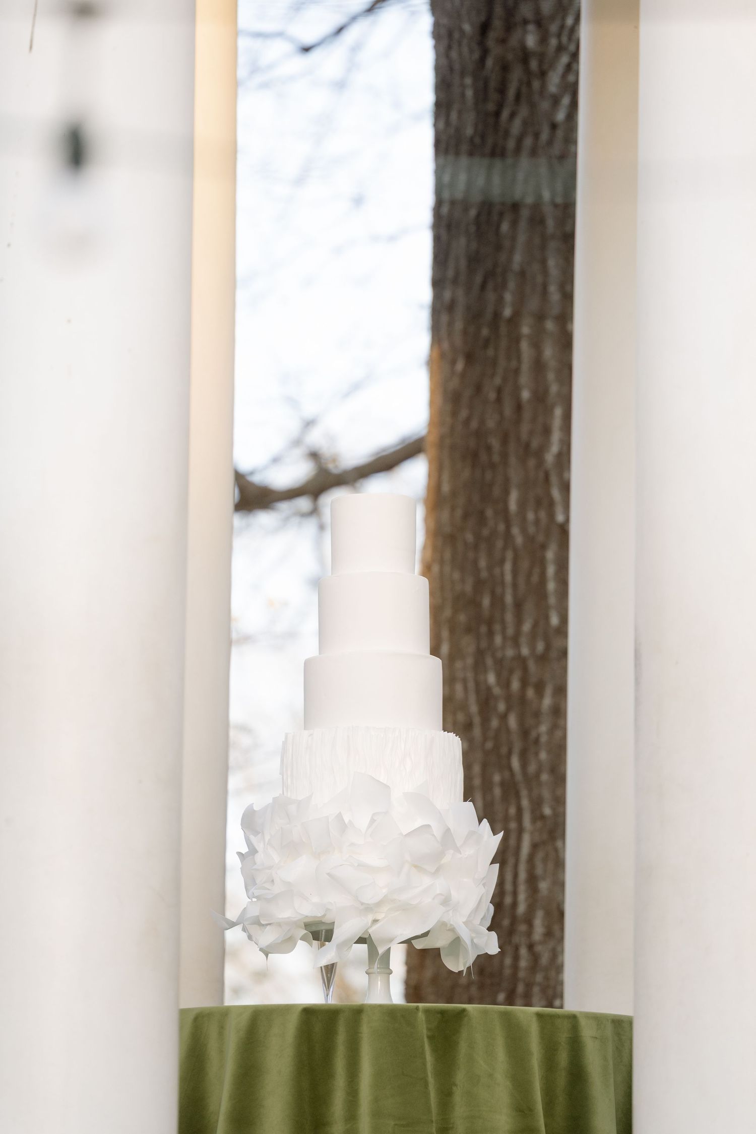 Elegant white multi-tiered wedding cake adorned with white floral details displayed on a green draped table near a window.