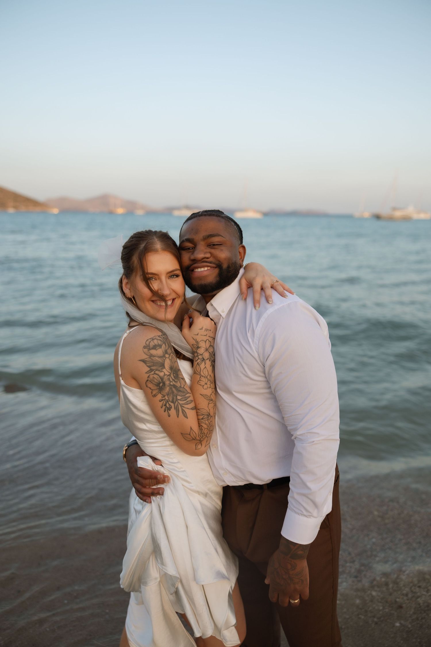 A romantic couple in white attire embraces on a beach at sunset with ocean waves in the background.