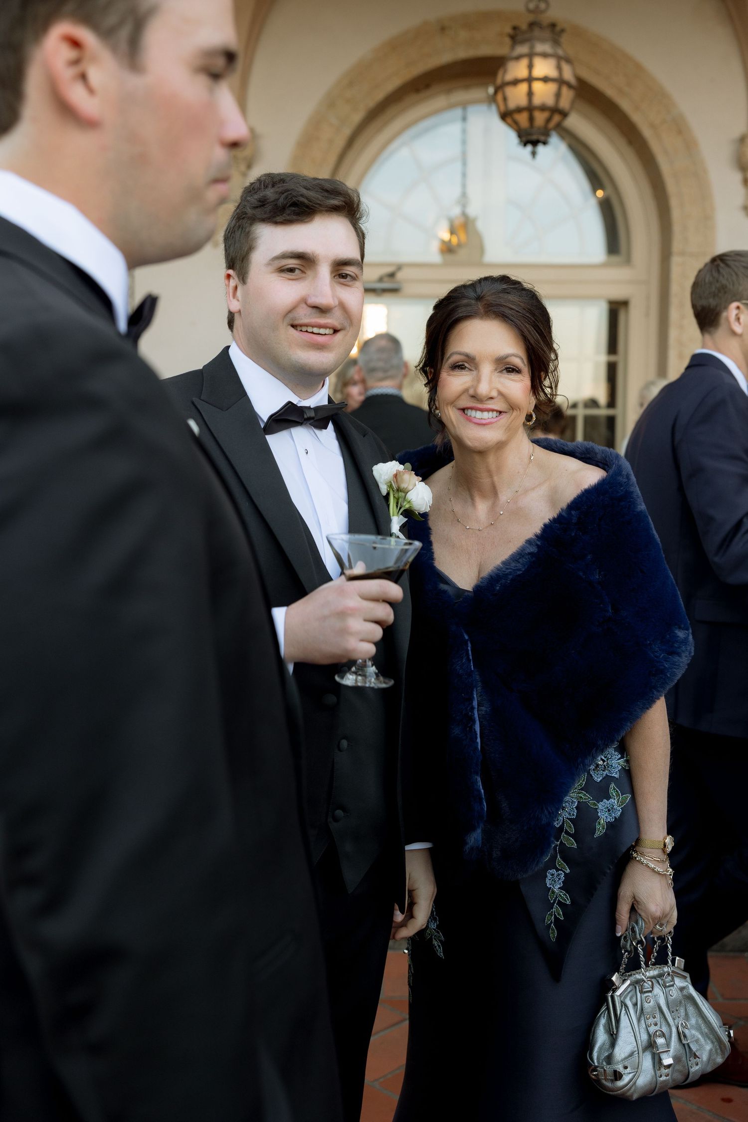 Groom in tuxedo poses with elegantly dressed woman in blue fur wrap at wedding reception.