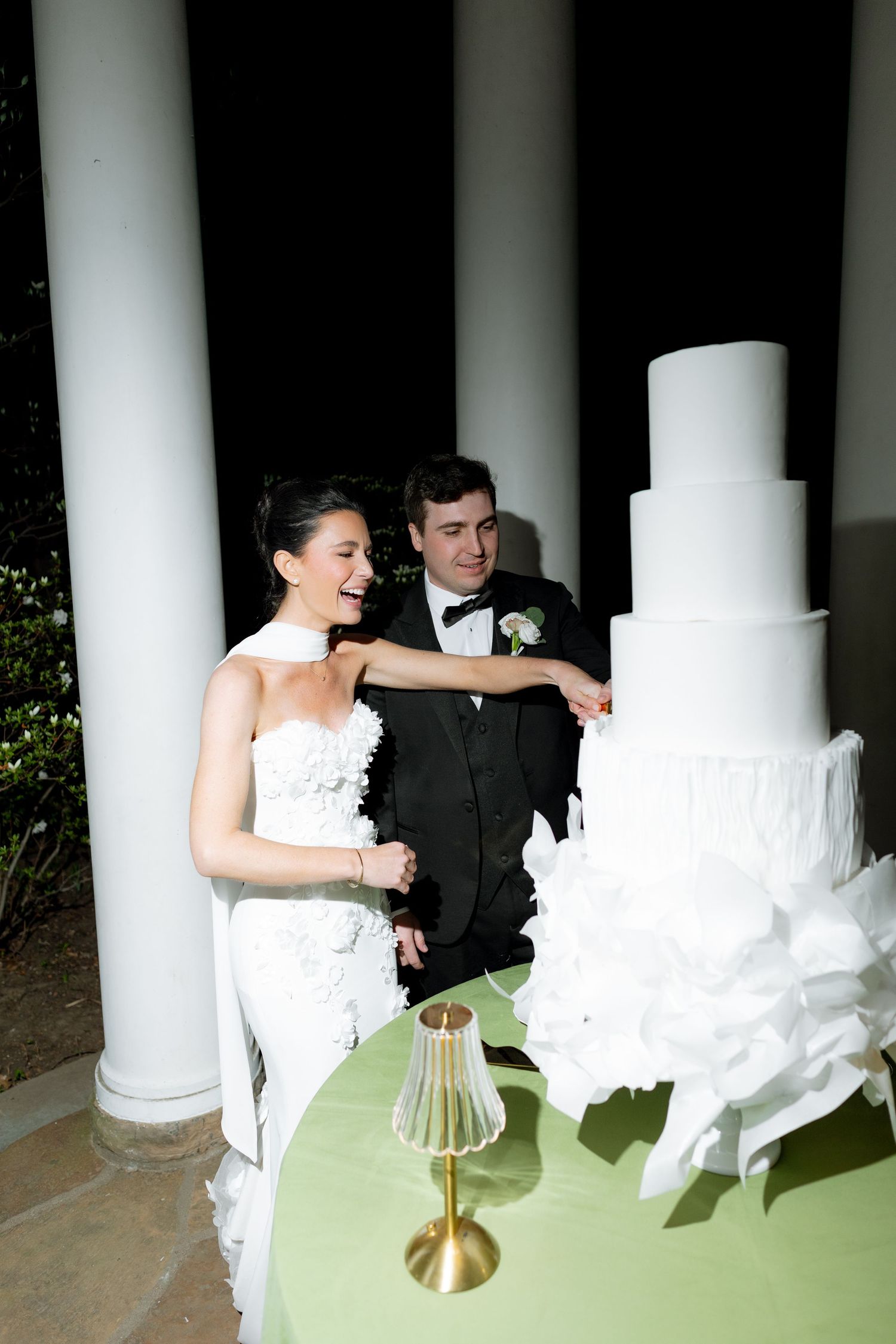 Smiling bride and groom cut elegant five-tier white wedding cake adorned with ruffles at night.