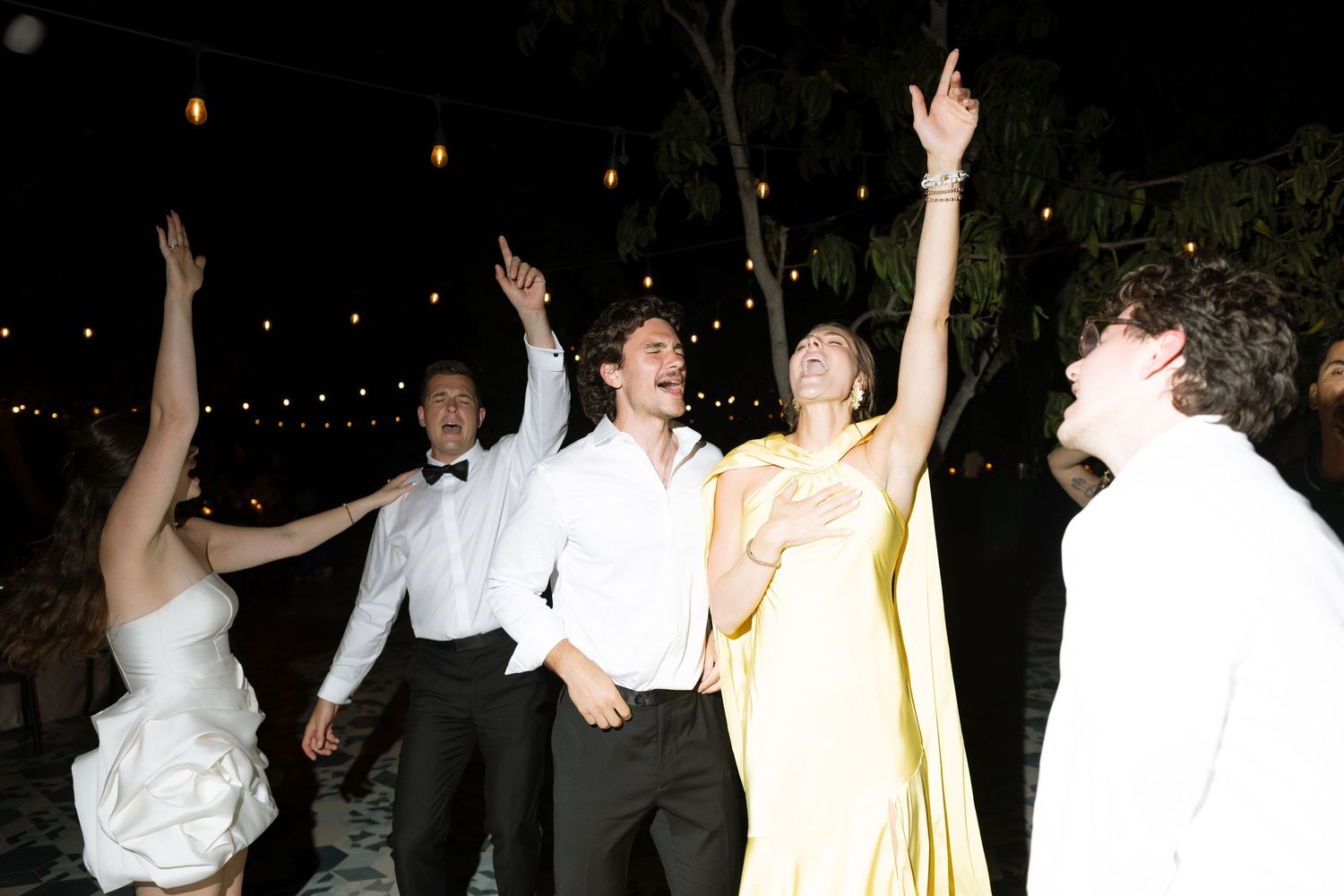 Wedding guests in formal attire dancing joyfully outdoors at night under string lights.