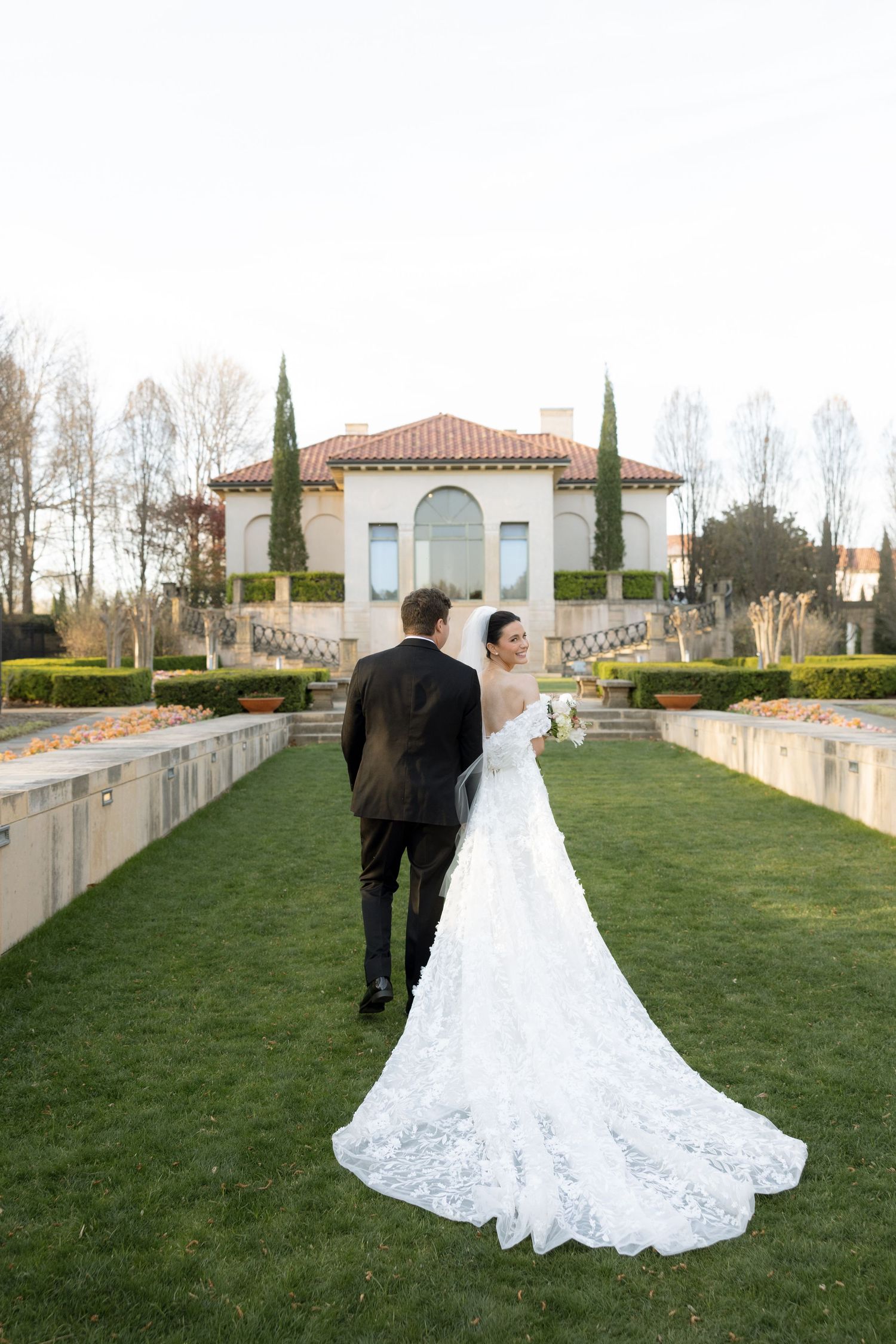 Bride and groom walk toward elegant Italian villa wedding venue with lush green lawn and manicured gardens.