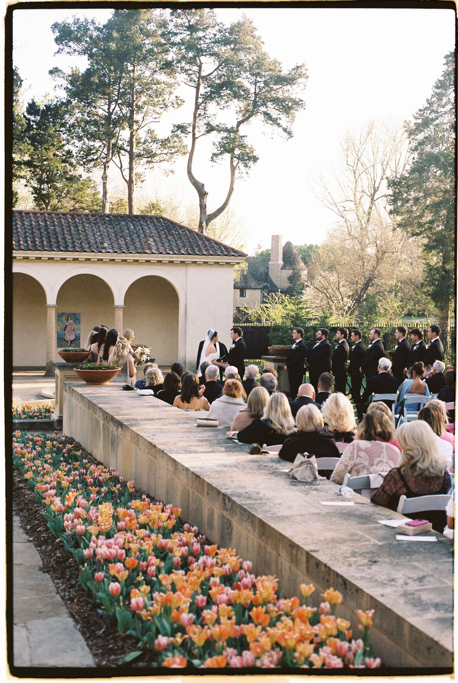 Outdoor wedding ceremony beside colorful tulip garden with guests seated and wedding party at altar near arched pavilion.