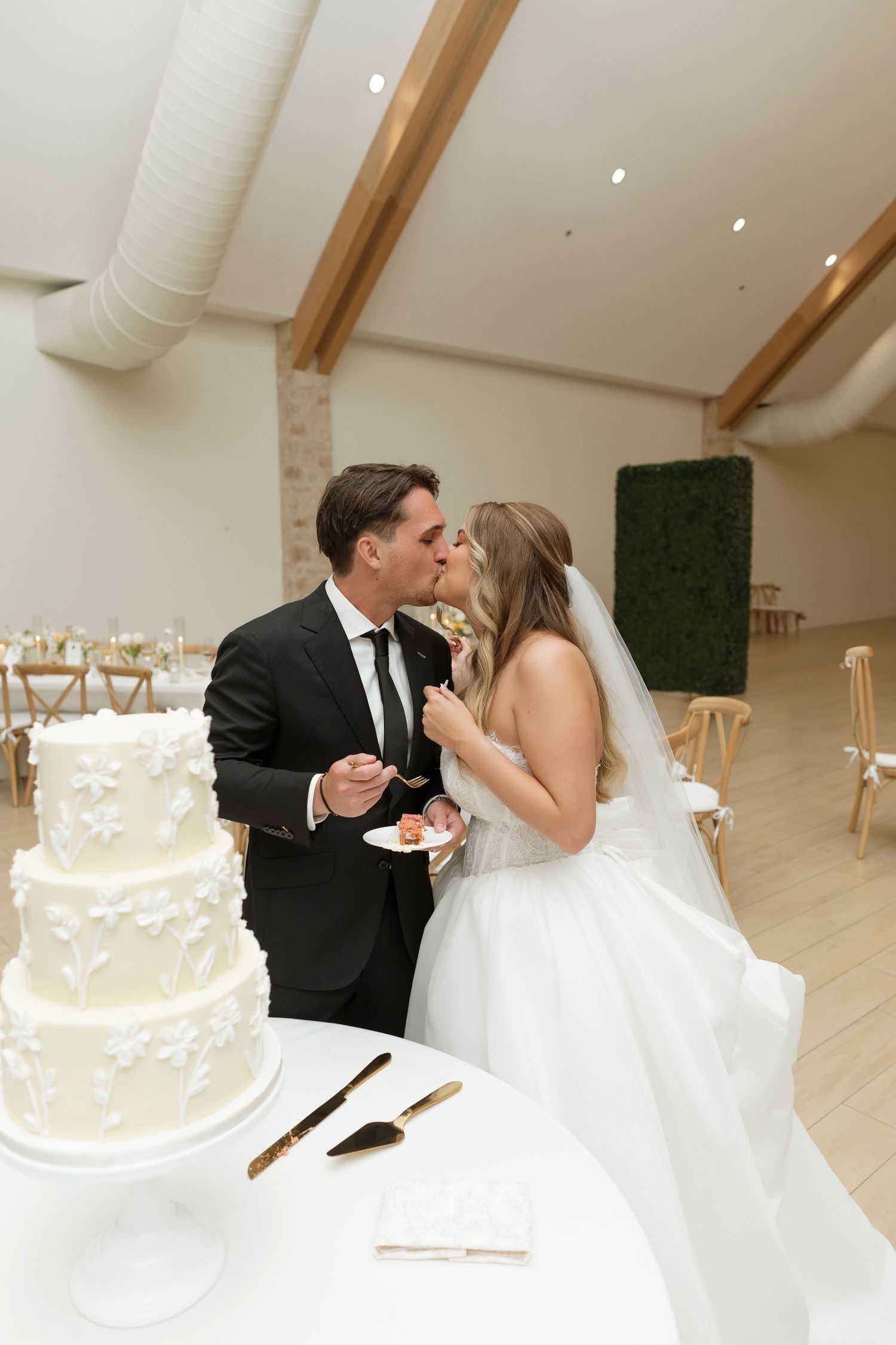 Newlyweds share a romantic moment near their elegant white wedding cake in a modern reception venue.