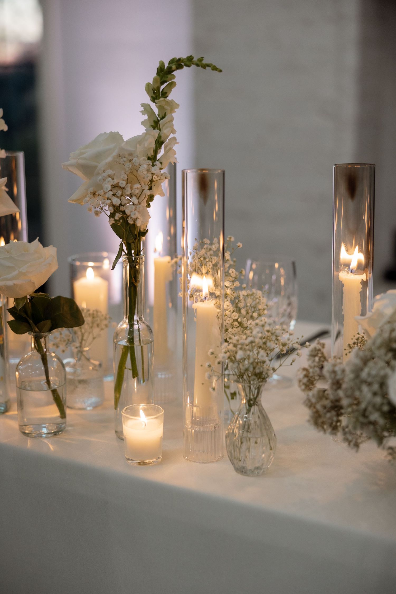 Romantic candlelit table setting with white flowers in glass vases and tall cylinder candle holders.
