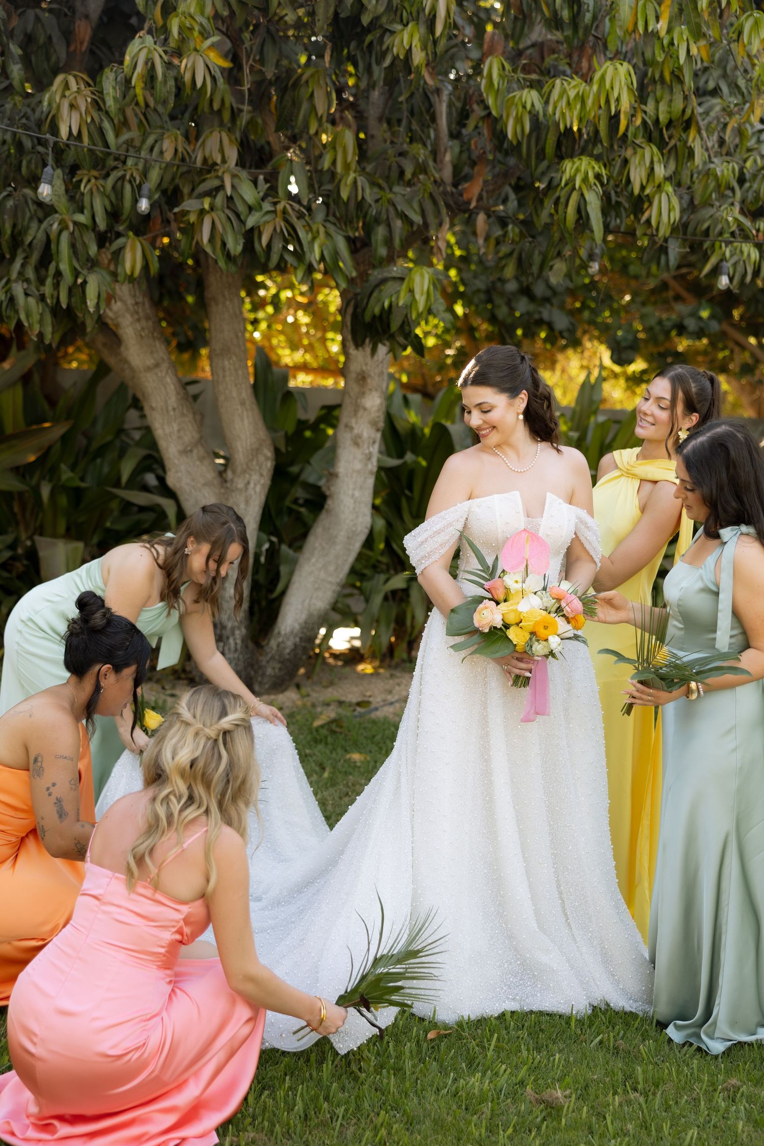 Bride in white off-shoulder gown smiles as bridesmaids in colorful dresses arrange her train outdoors.