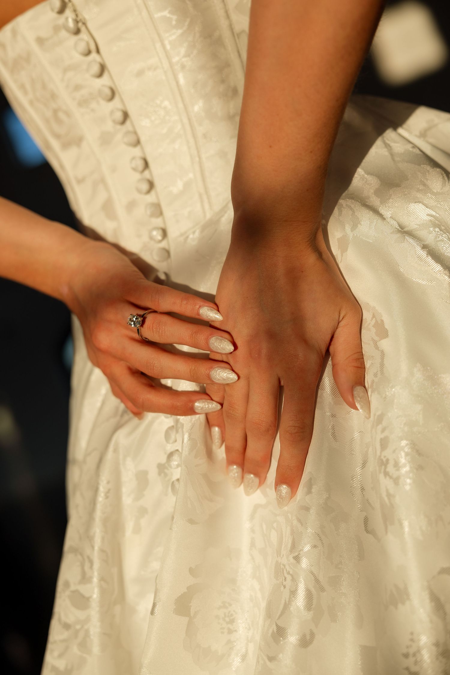 Close-up of hands wearing engagement ring resting on white lace wedding dress fabric.